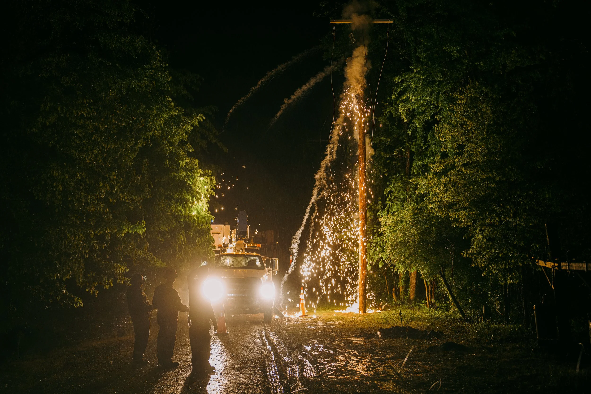 Power line sparking during nighttime as utility workers stand near a truck with headlights on, surrounded by trees.