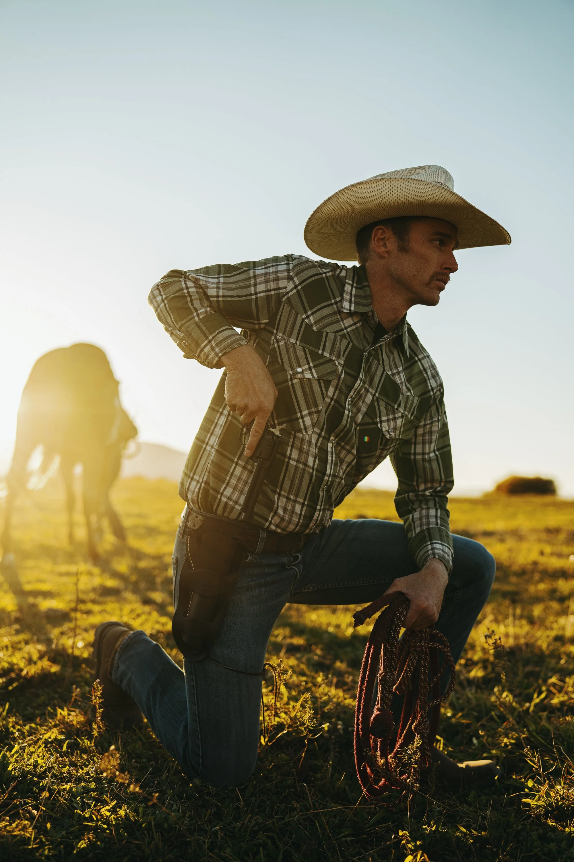 Cowboy kneeling on grass with horse in the background, wearing a plaid shirt and cowboy hat, holding a revolver in a holster and coiled rope.