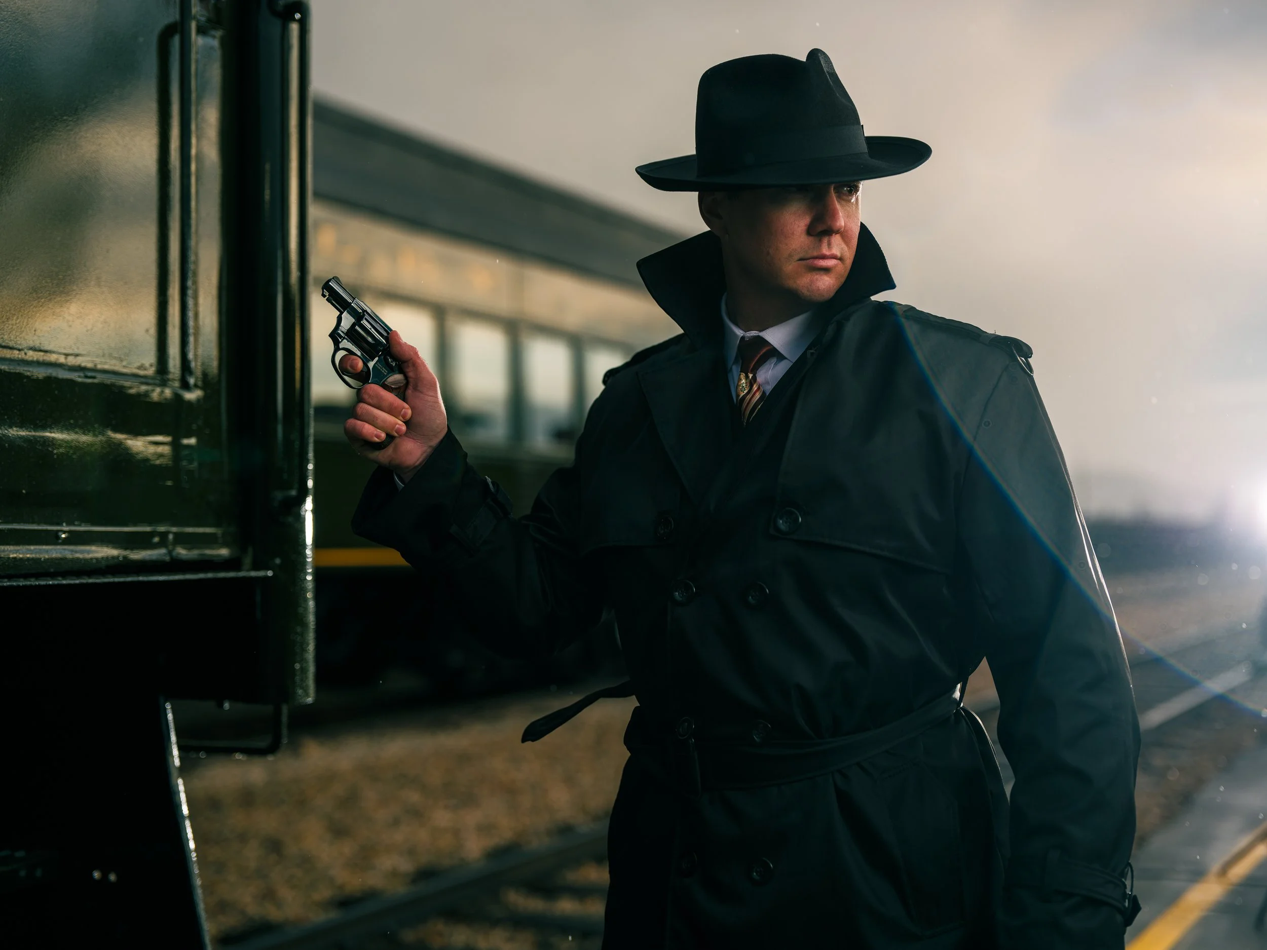 Man in trench coat and fedora holding a revolver near a train, with a dramatic and moody atmosphere.