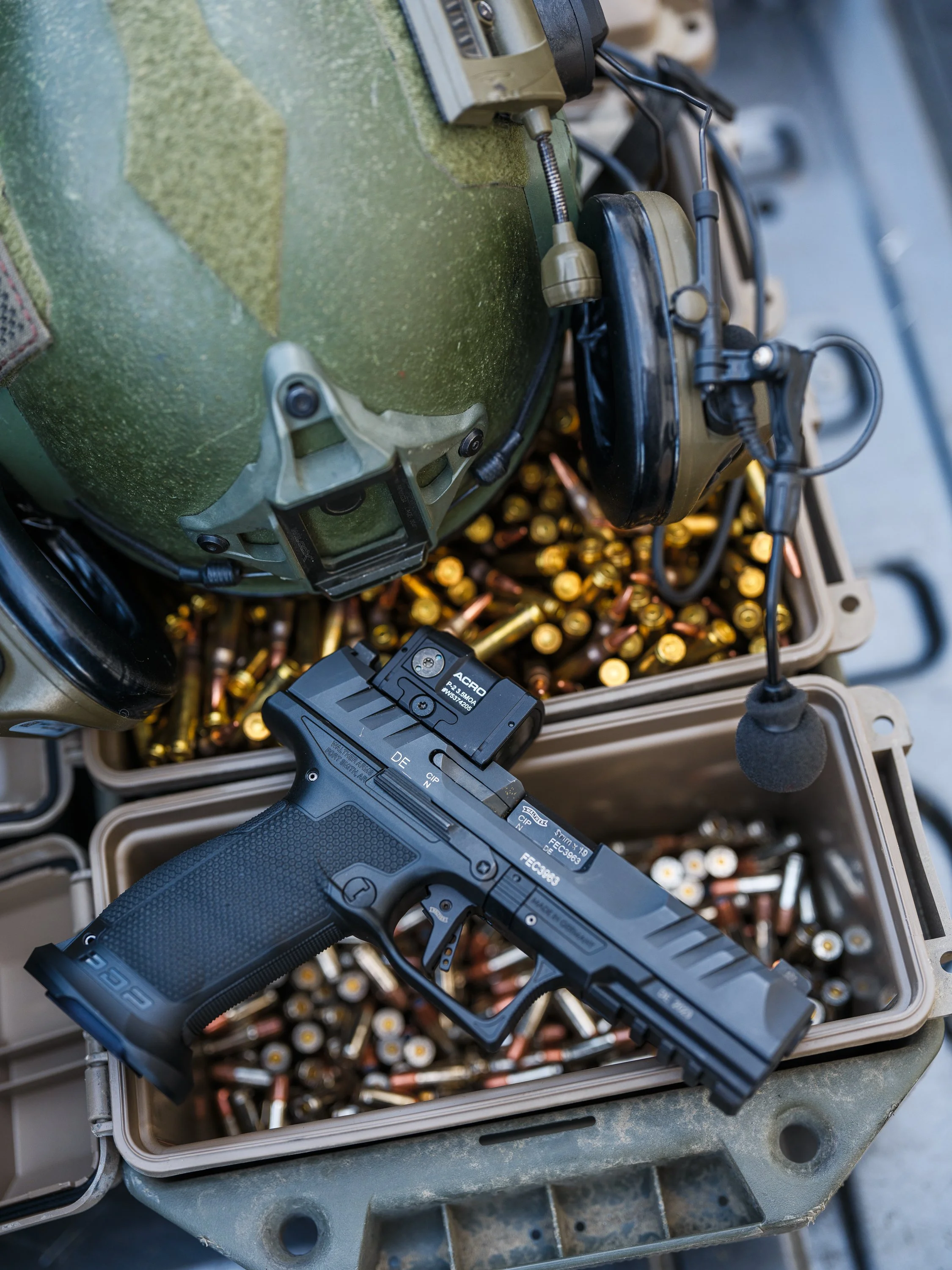 Black handgun with tactical sight on a box of ammunition beside a green military helmet with attached headset.