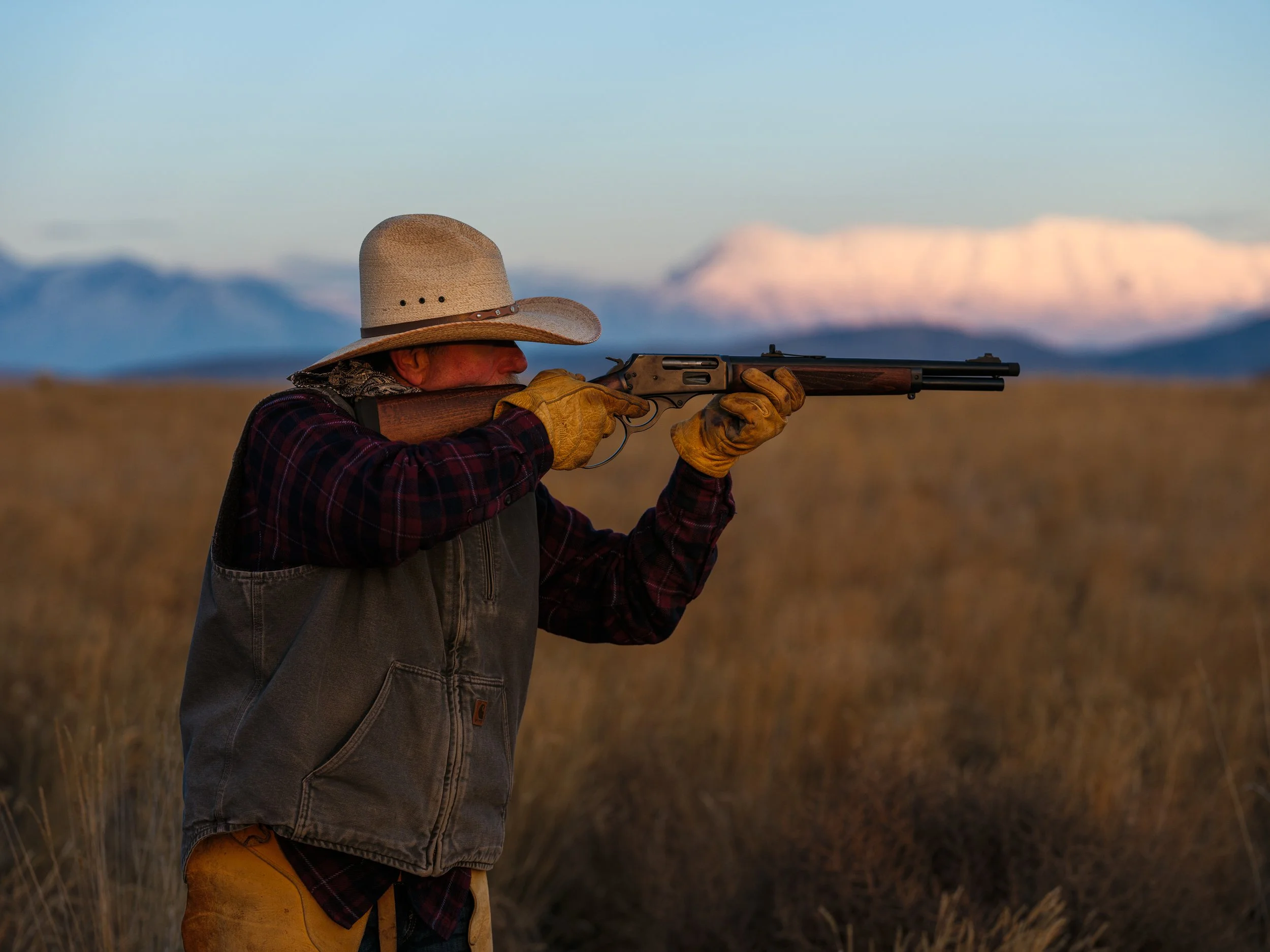 Man in cowboy hat aiming a rifle in a field with mountains in the background at sunset.