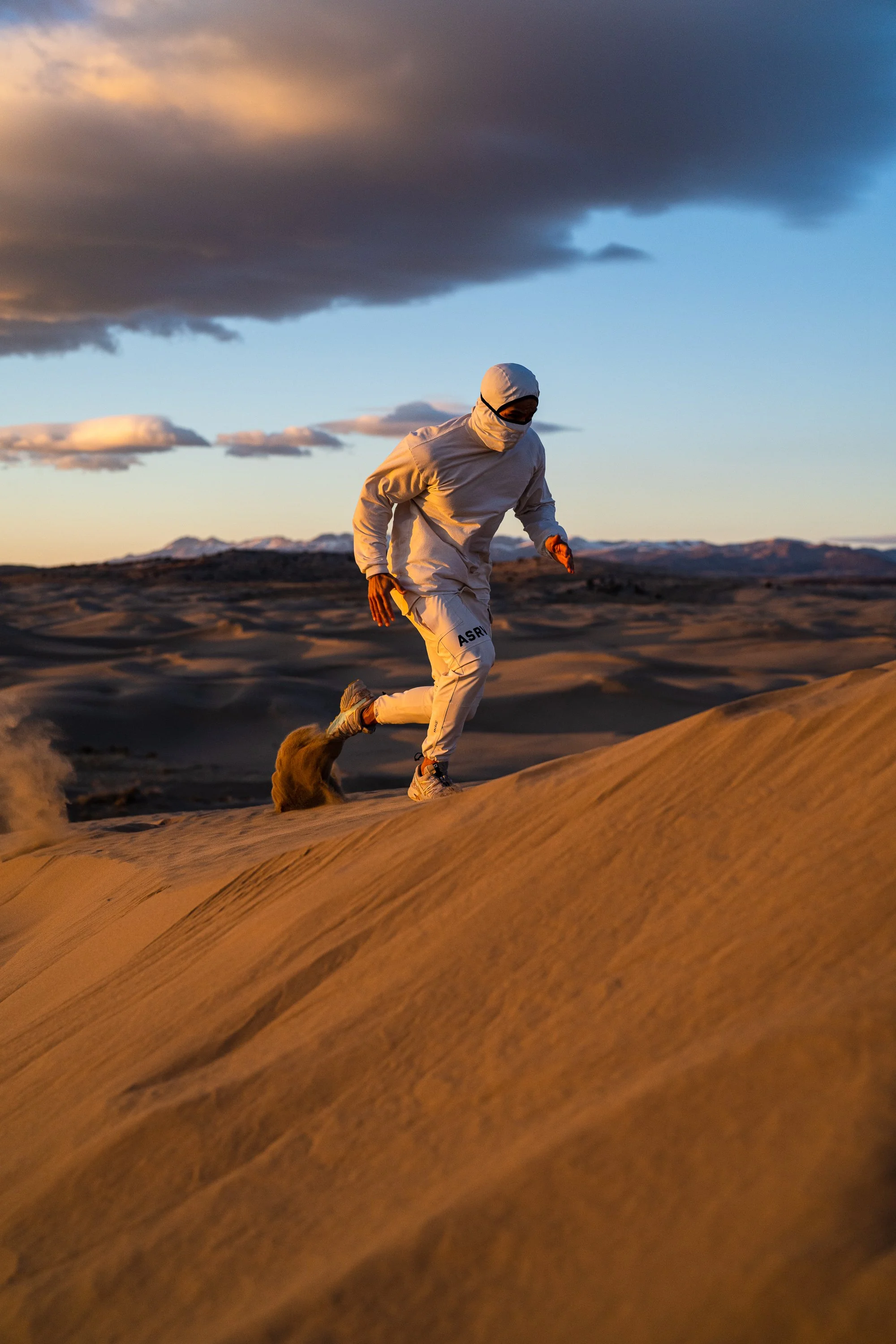 Person wearing white athletic gear running on sand dune during sunset with dramatic clouds in the sky.