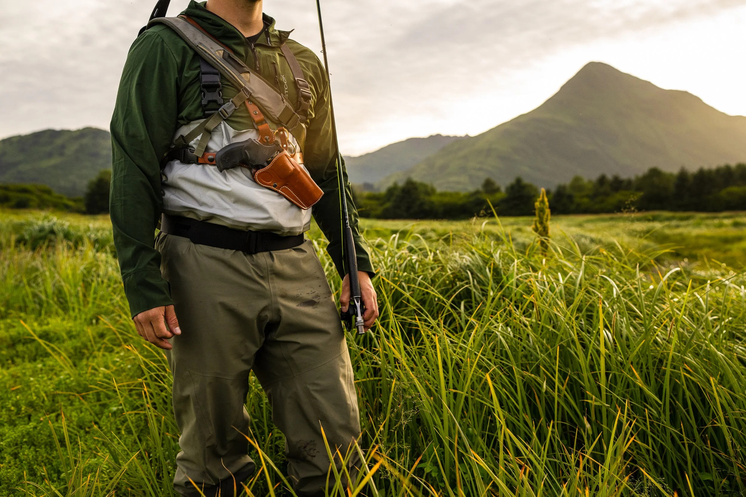 Person in outdoor gear with fishing rod standing in tall grass, mountain in background.