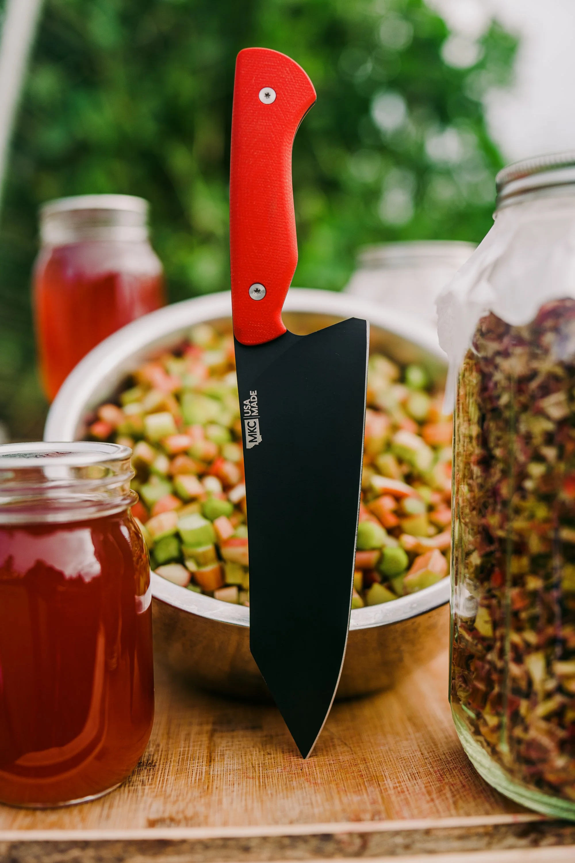 Red-handled kitchen knife, metal bowl with chopped vegetables, jars with liquids on a wooden surface, outdoor setting.