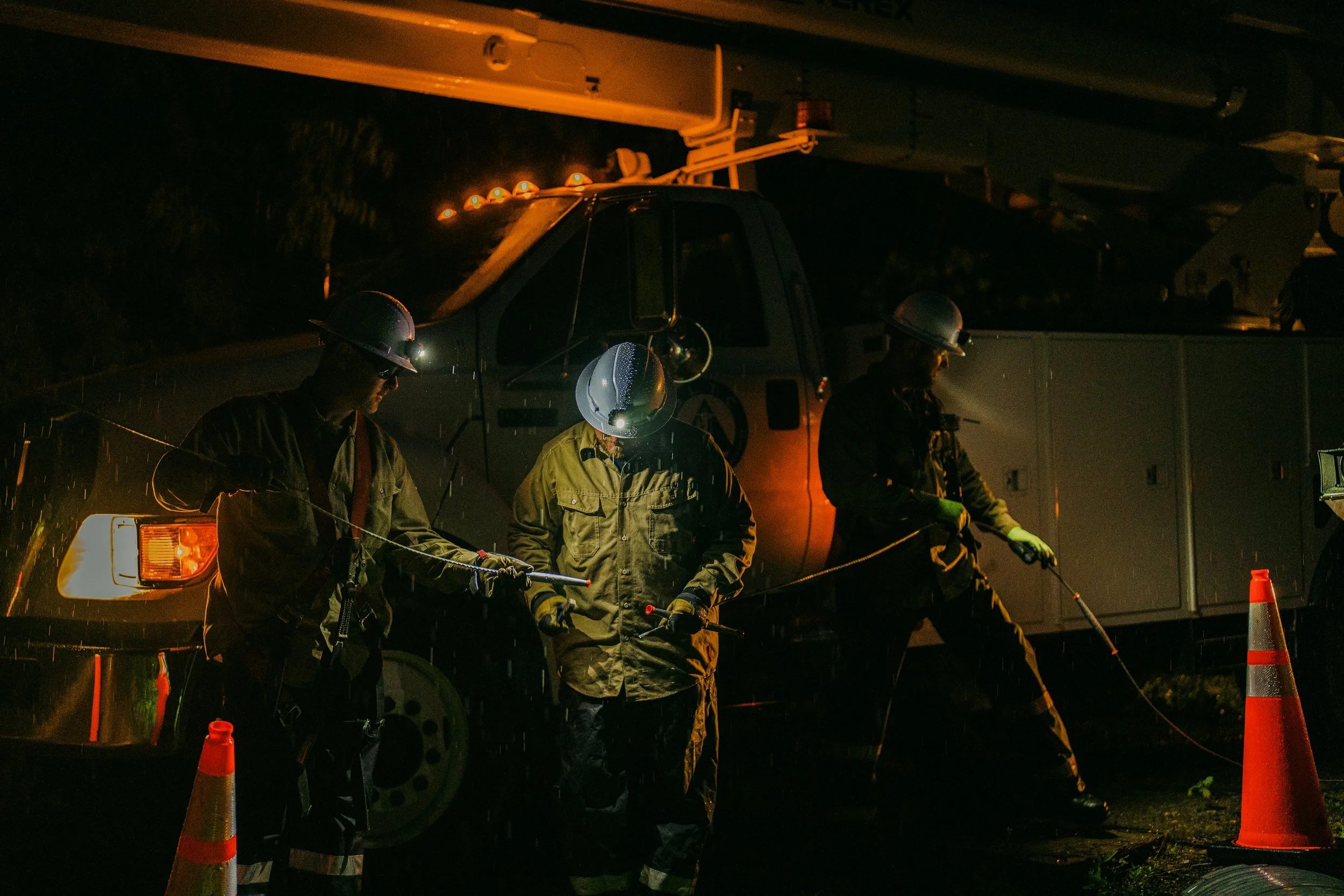 Three construction workers in hard hats and reflective gear working at night near a utility truck with a cone, illuminated by vehicle lights.