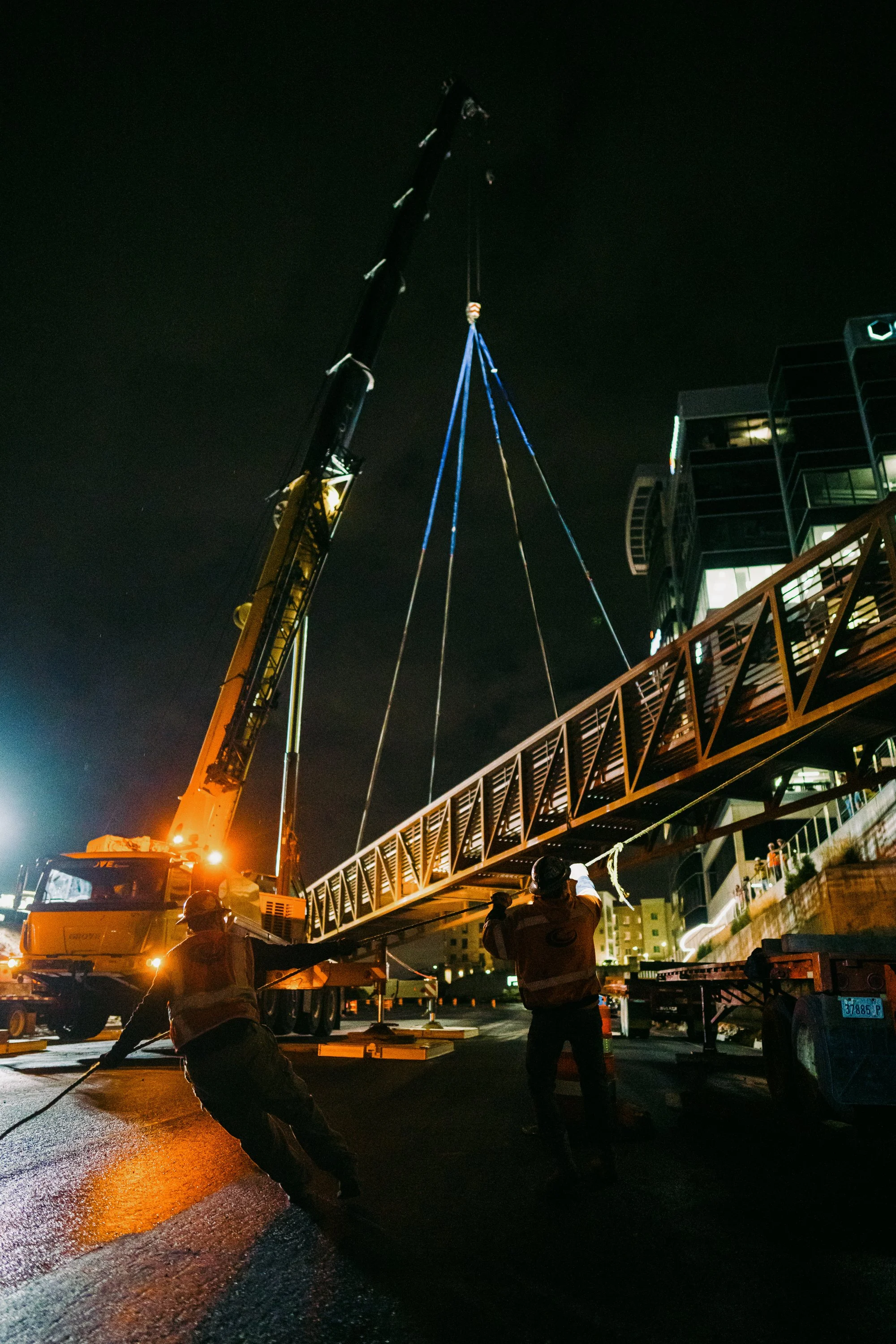 Crane lifting a pedestrian bridge at night with workers guiding the process.
