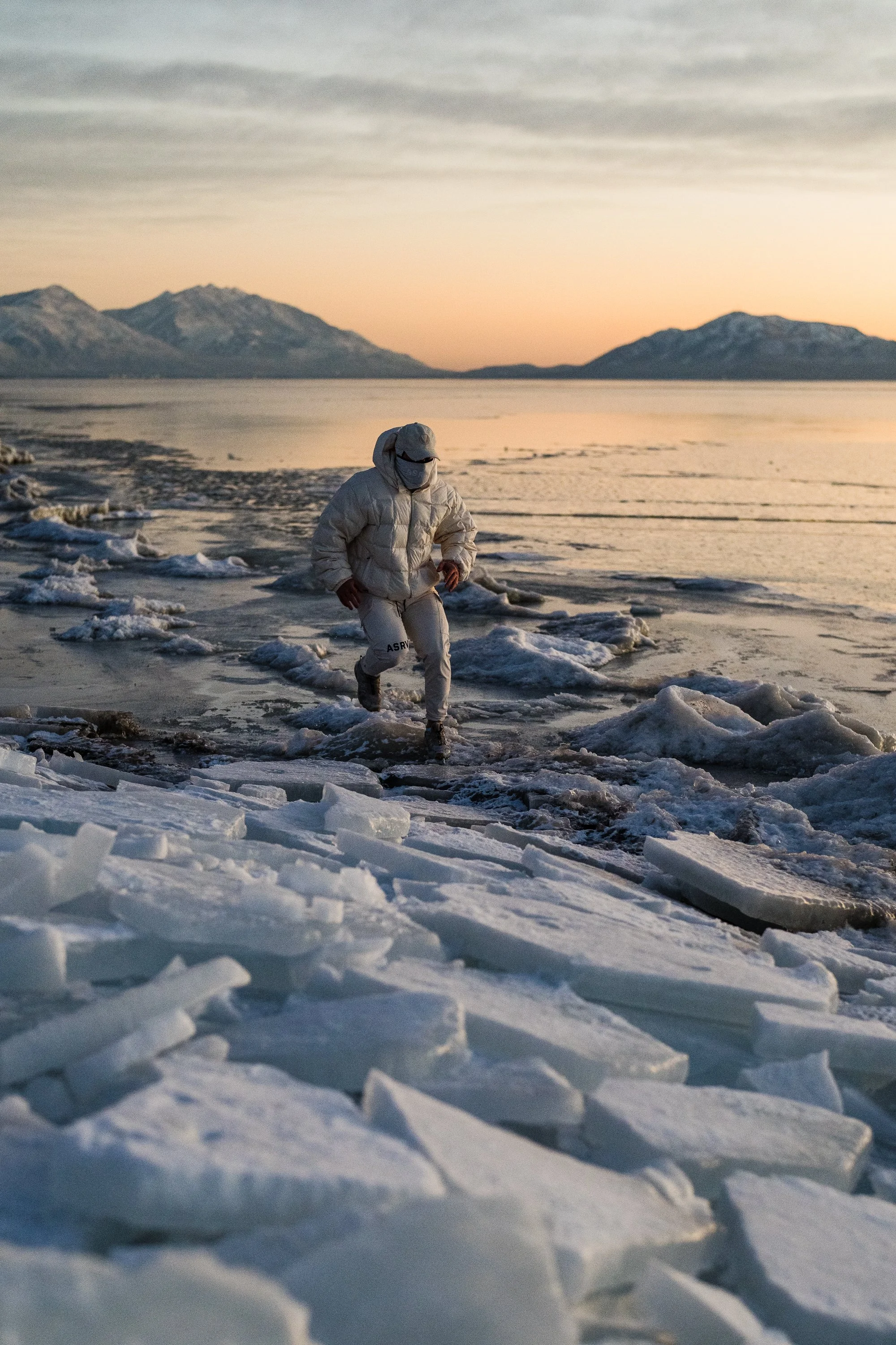 Person in white clothing walking on icy terrain by a frozen lake with snowy mountains in the background at sunset.