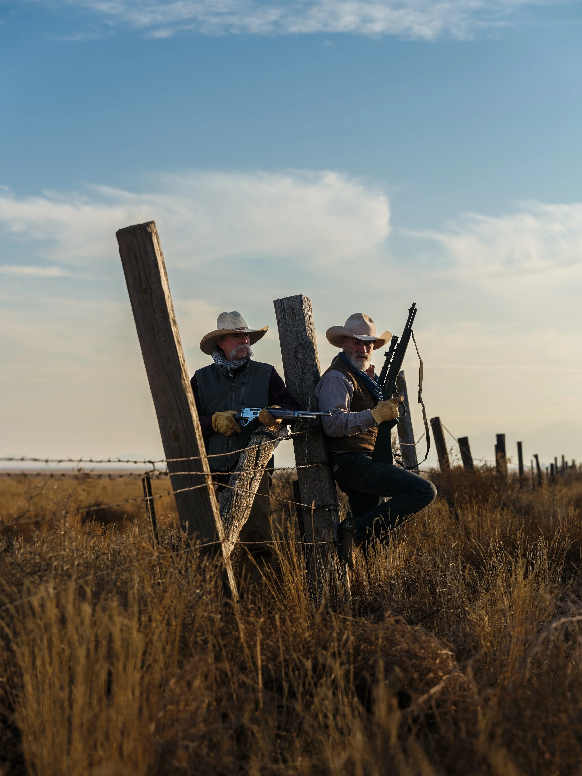Two cowboys in hats and gloves leaning against wooden fence posts, each holding rifles, with a rural landscape and barbed wire. The setting is outdoors, under a clear blue sky.