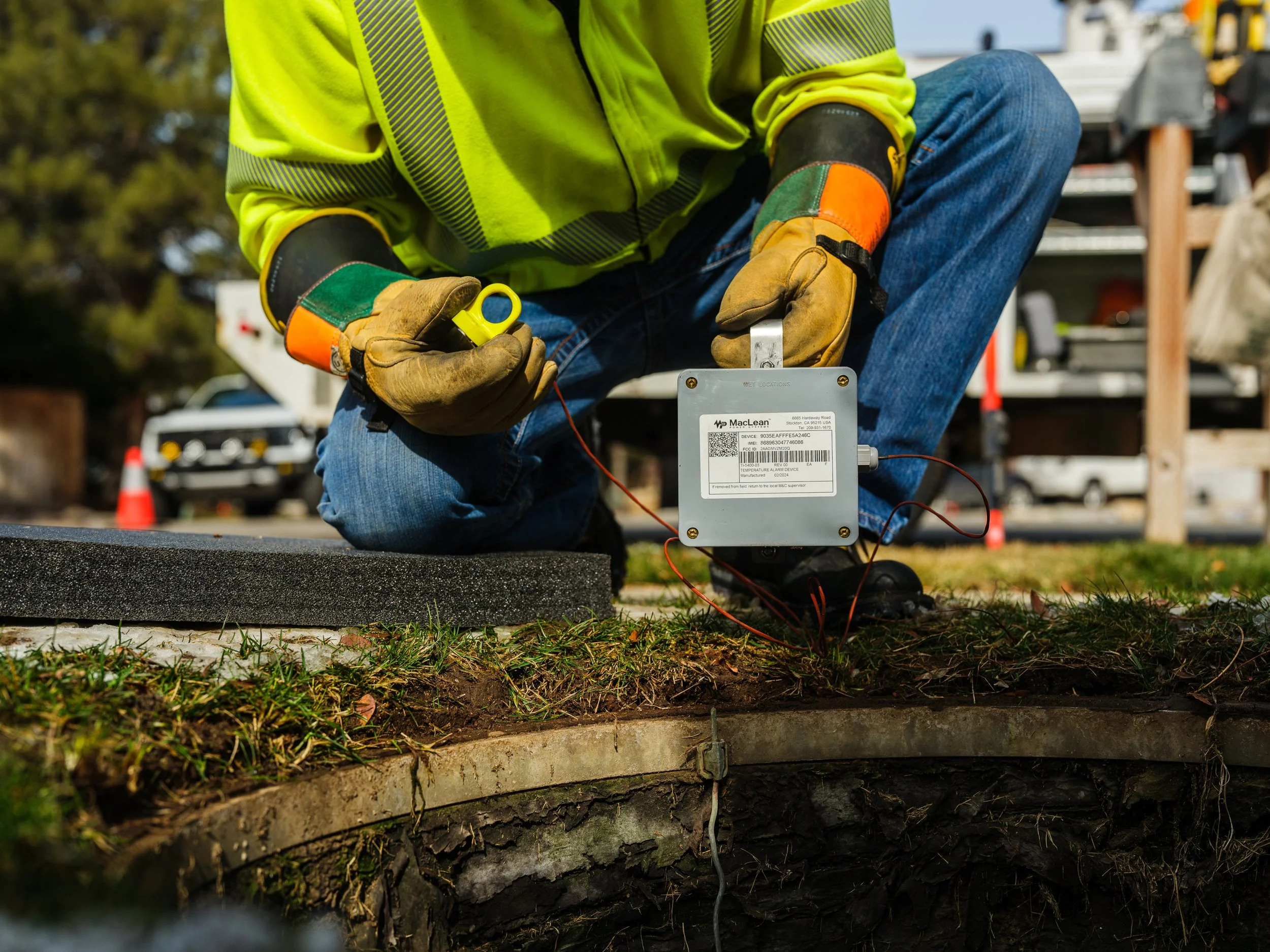 A worker wearing protective gloves and a neon yellow safety jacket installs a device near a manhole on the grass. The device is connected by wires, and a vehicle and traffic cone are visible in the background.