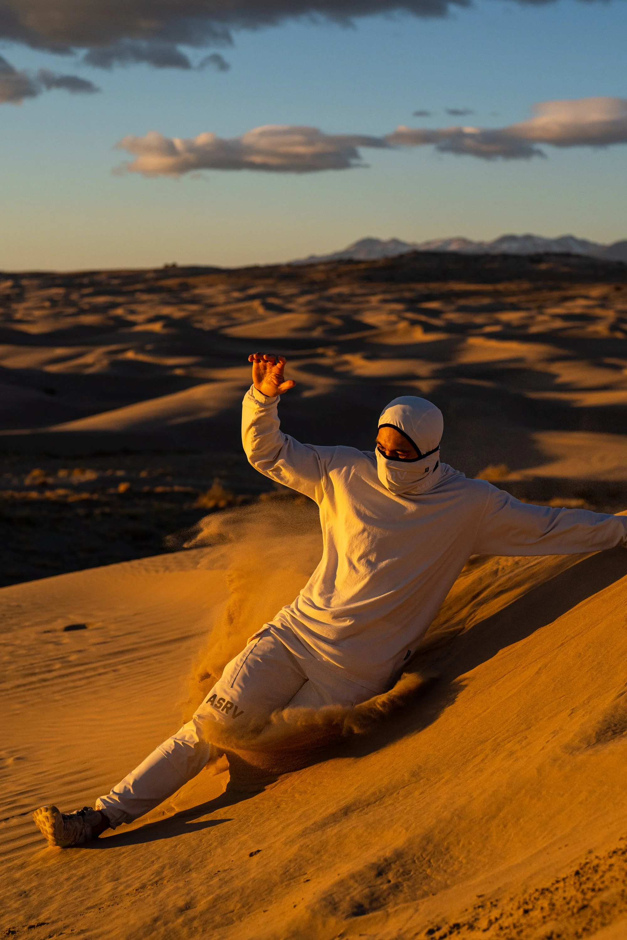 Person sandboarding in desert landscape at sunset, wearing white clothing and mask.