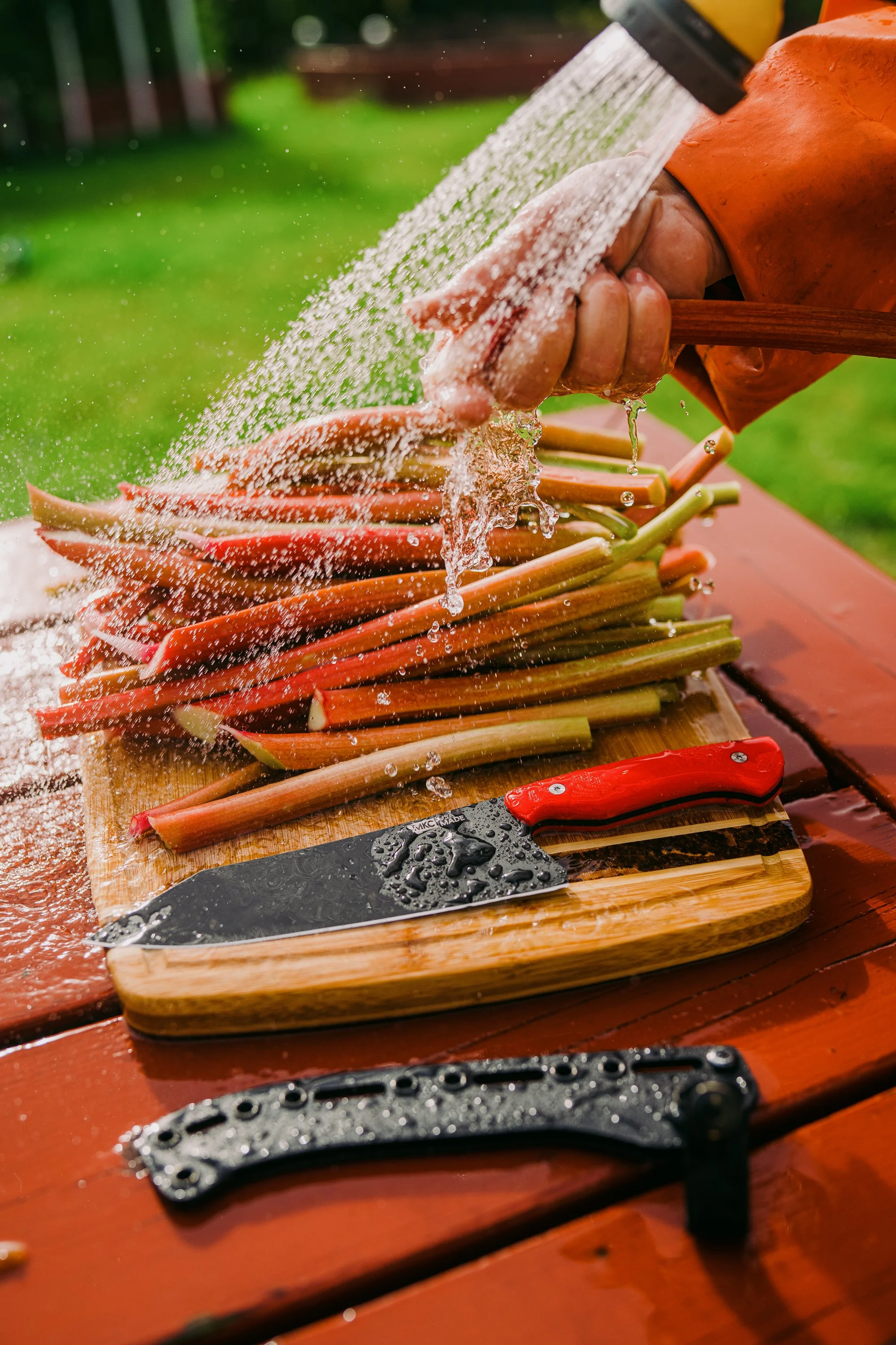 Person washing rhubarb stalks with water on a wooden cutting board next to a knife with a red handle.