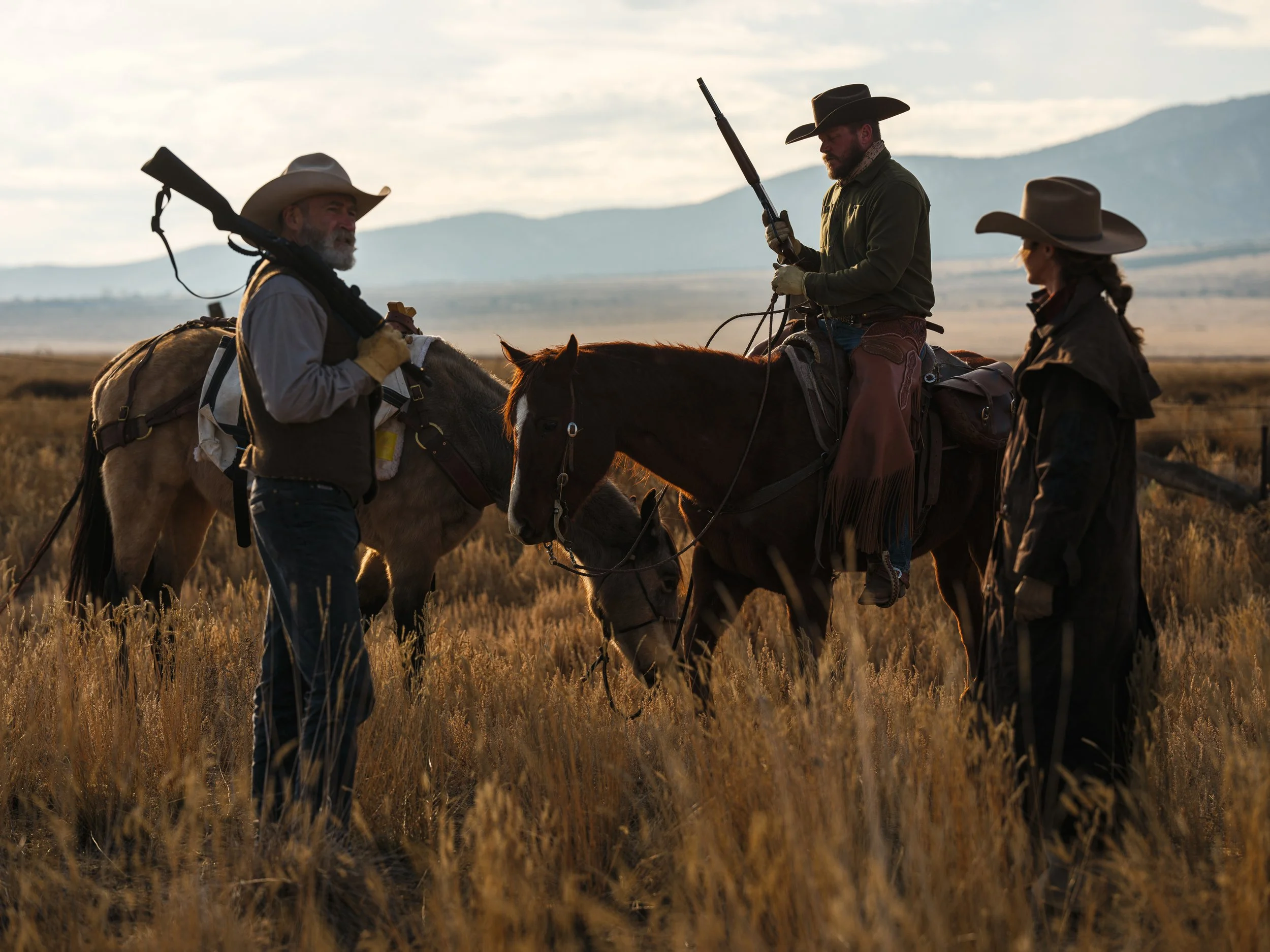 Three cowboys in a grassy field, two on horseback, holding rifles, with mountains in the background.