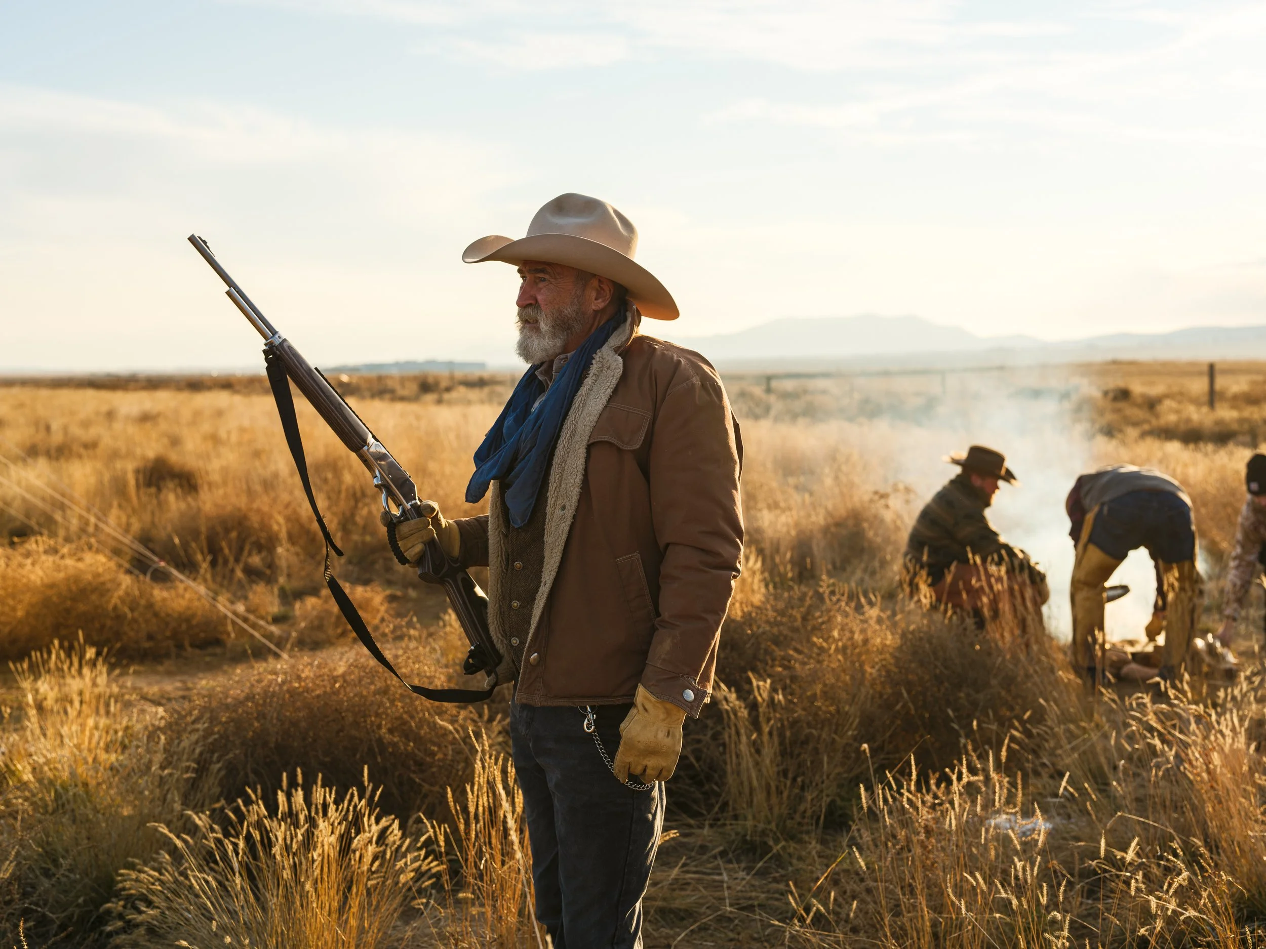 Man in cowboy attire holding a rifle in a dry grassland, with two others tending a campfire in the background.
