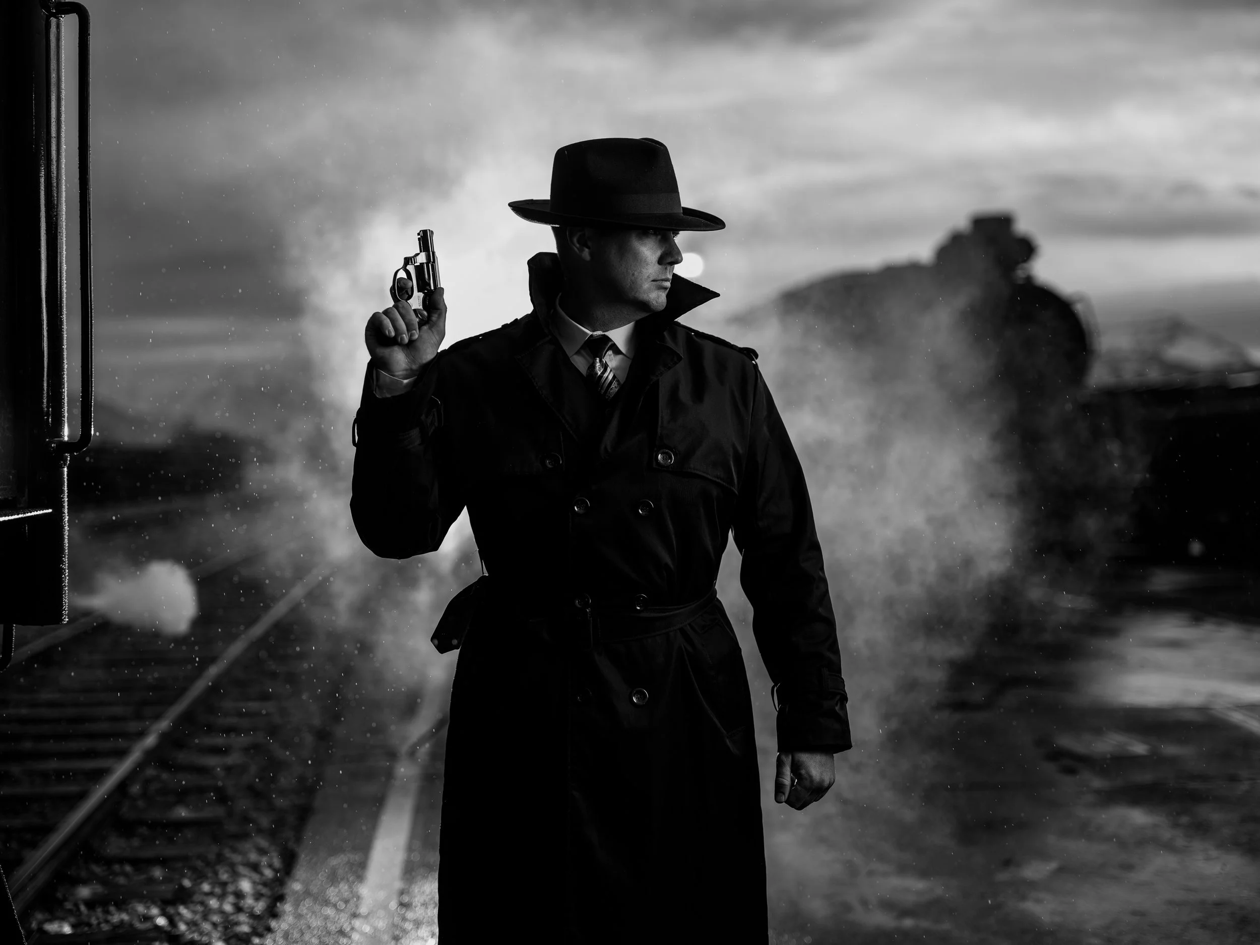 Black and white photo of a man in a trench coat and fedora hat holding a revolver with a steam engine in the background, evoking a classic noir atmosphere.