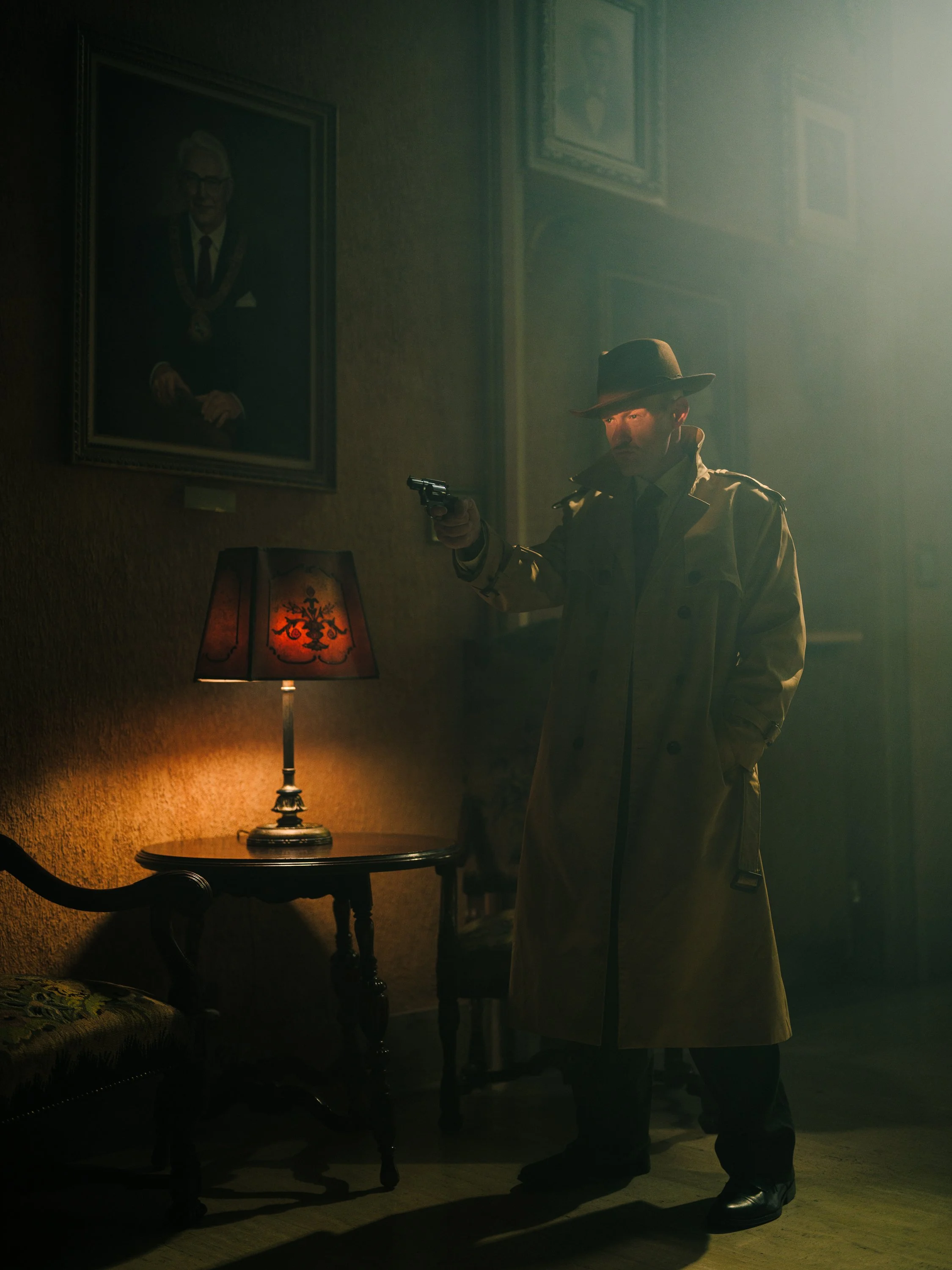 A detective in a trench coat and fedora hat holding a revolver in a dimly lit room. Beside him is a small table with a lamp, an armchair, and portraits on the wall.