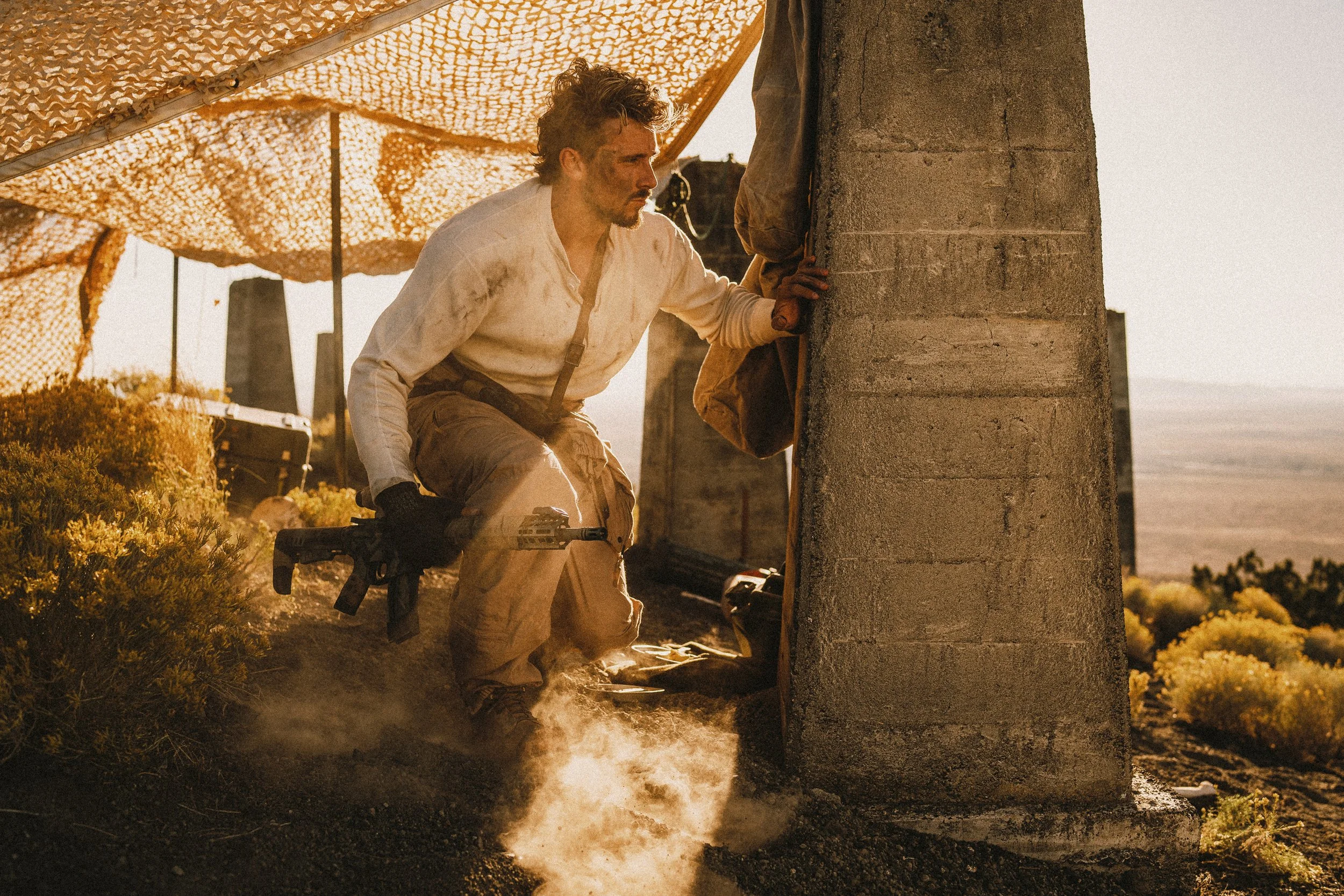 A man wearing a dirty white shirt and khaki pants crouches beside a concrete pillar, holding a tool or weapon. He is under a camouflage net in a dry, rocky landscape with scattered bushes. Sunlight casts strong shadows, creating a dramatic effect.