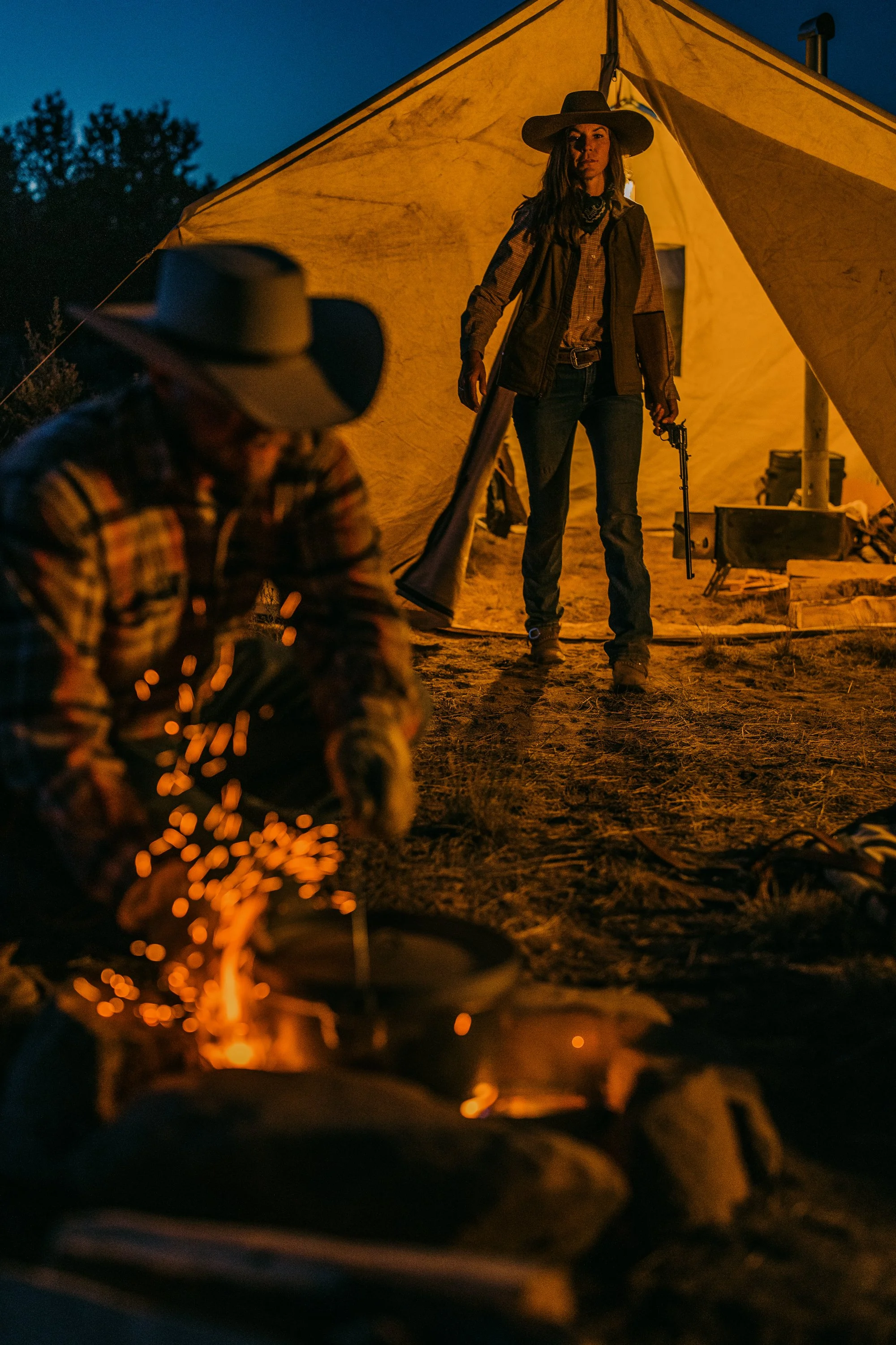 Two cowboys outdoors at night, one tending a campfire with sparks flying, the other standing near a canvas tent holding a rifle, wearing cowboy hats.