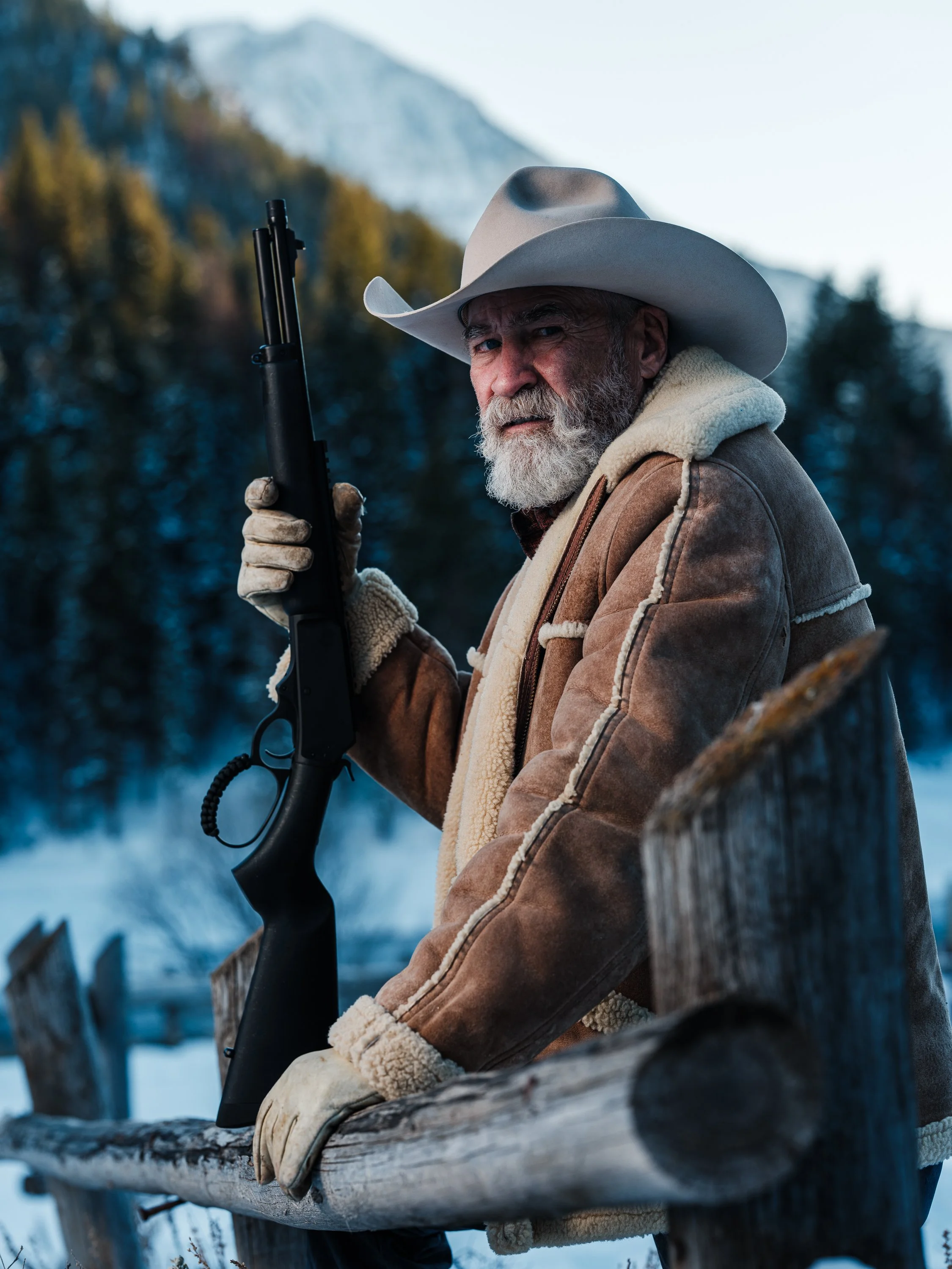 Man in a cowboy hat and fur-lined coat holding a rifle against a snowy mountain backdrop.