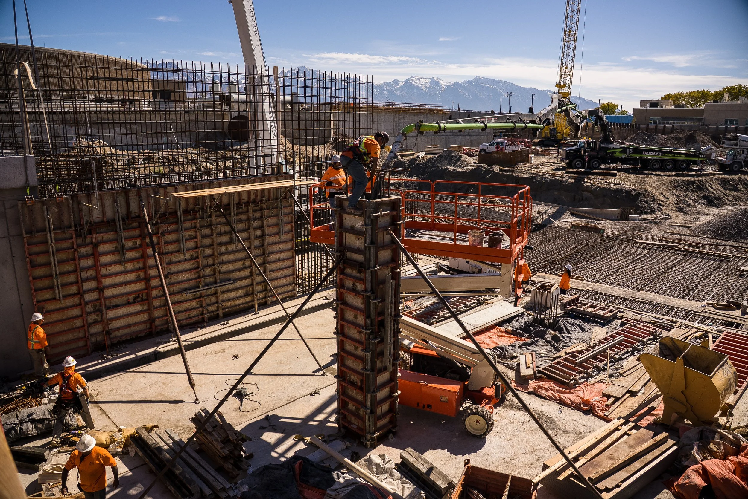 Construction site with workers and equipment, including cranes and rebar, set against a backdrop of mountains.