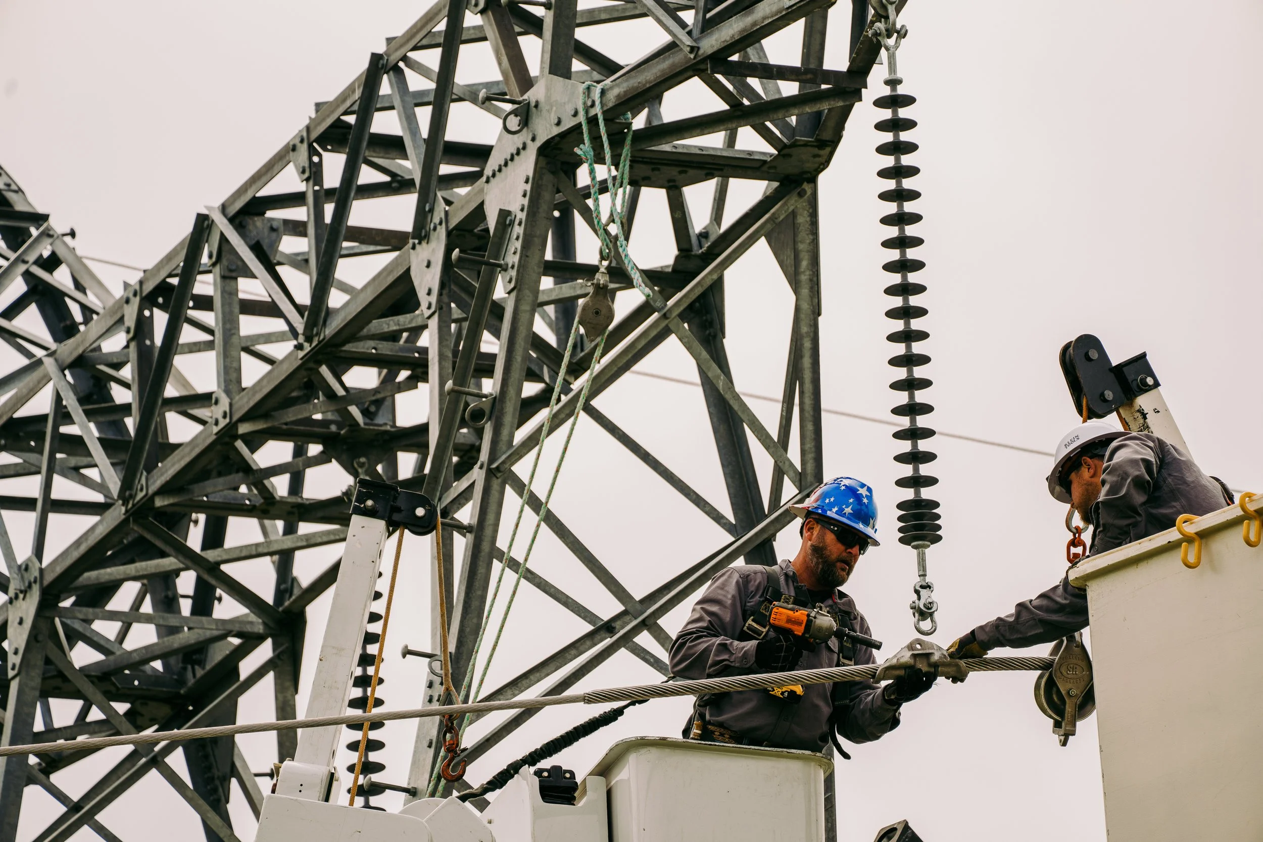 Two workers in hard hats and safety gear on a utility lift, maintaining a transmission tower.