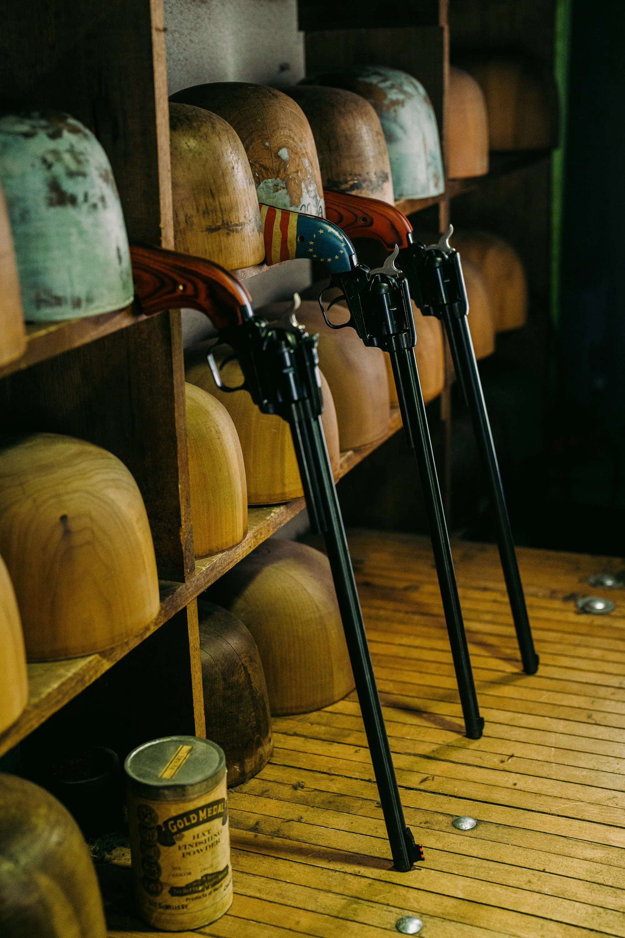 Three vintage revolvers with long barrels and American flag designs lean against a shelf with wooden head forms. A vintage tin container is on the wooden floor.
