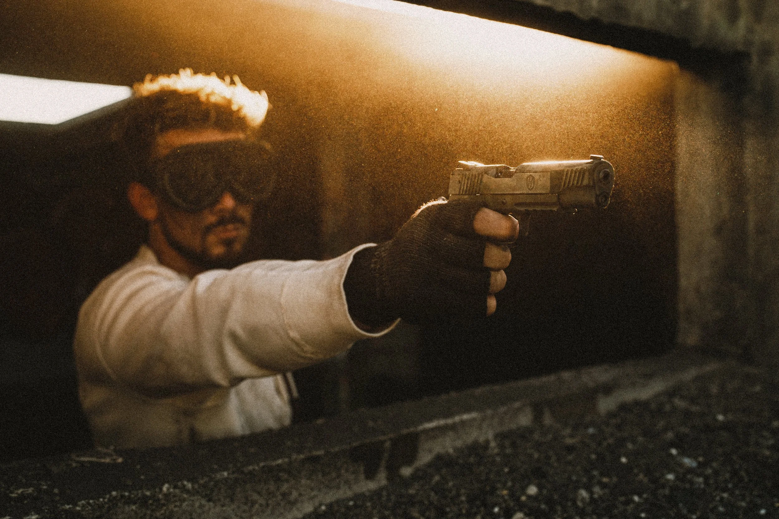 Man wearing goggles holding a handgun in a dimly lit environment with sunlight streaming in.
