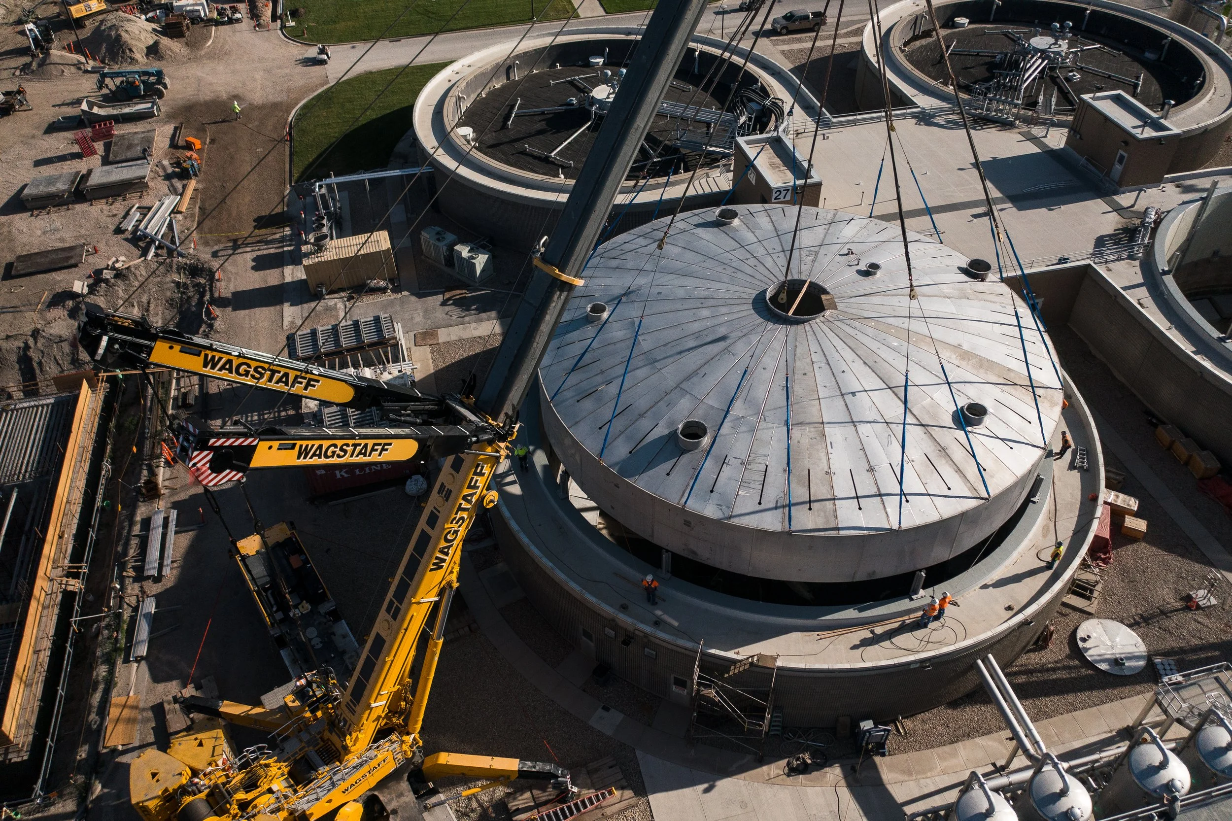 Aerial view of construction site with large circular industrial tanks, cranes labeled "Wagstaff," and workers in hard hats.