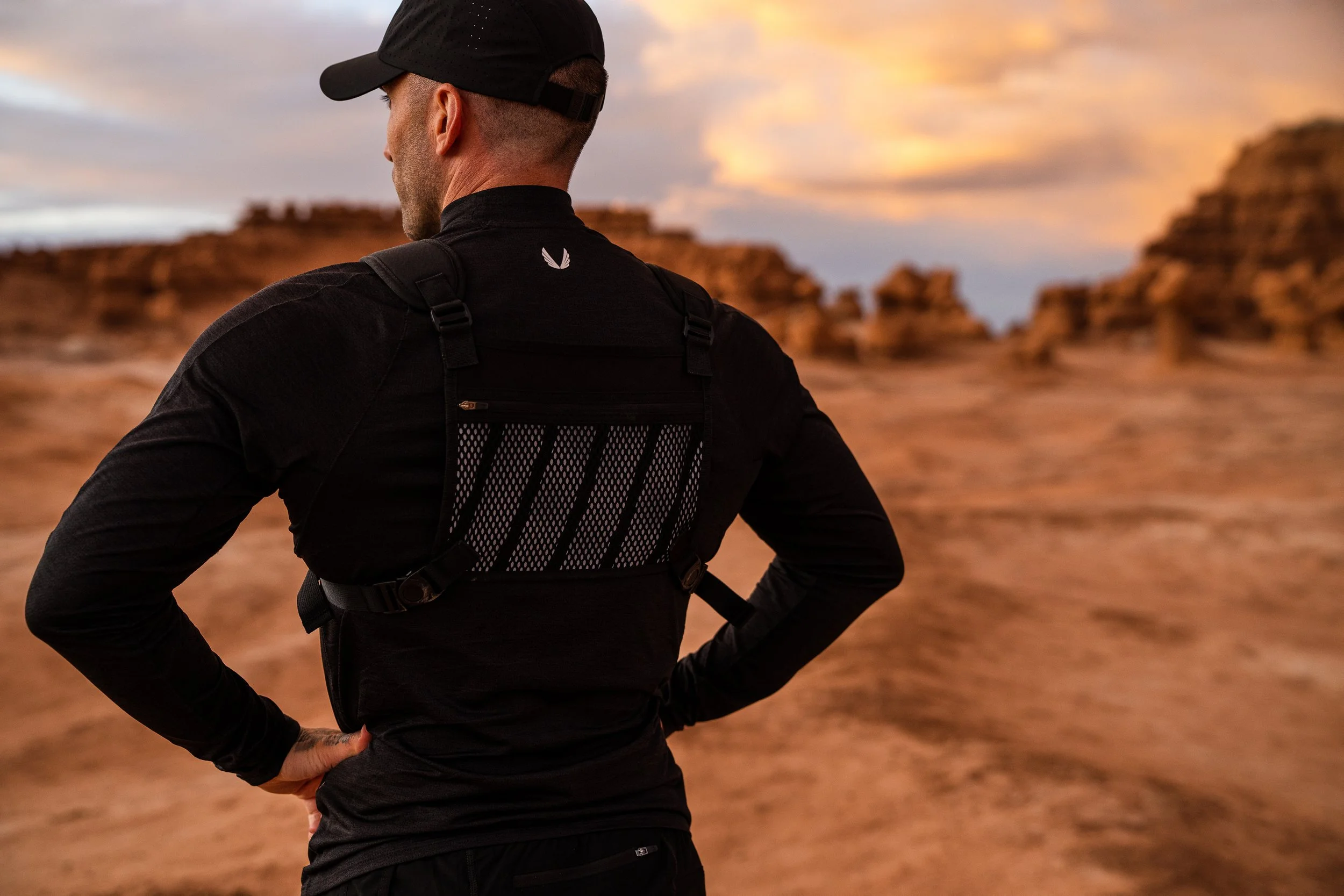 A person wearing a black shirt and cap with a tactical vest stands in a desert landscape during sunset.