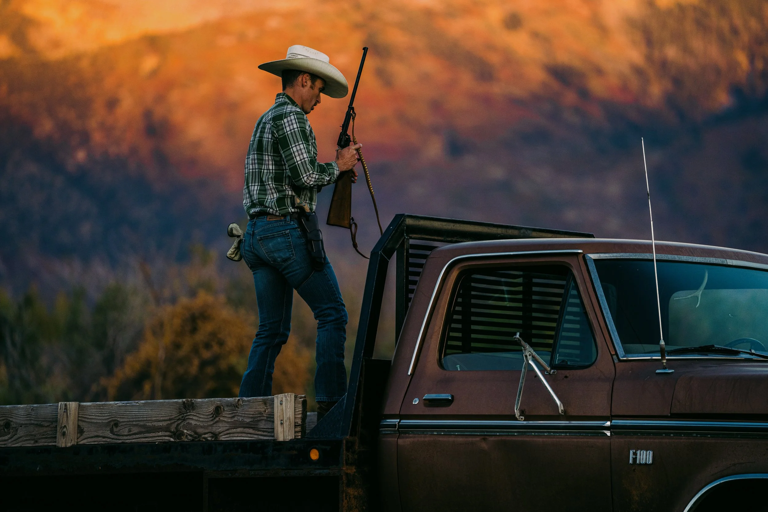 Person in cowboy hat and plaid shirt holding a rifle, standing on a flatbed truck, with a scenic mountain backdrop.