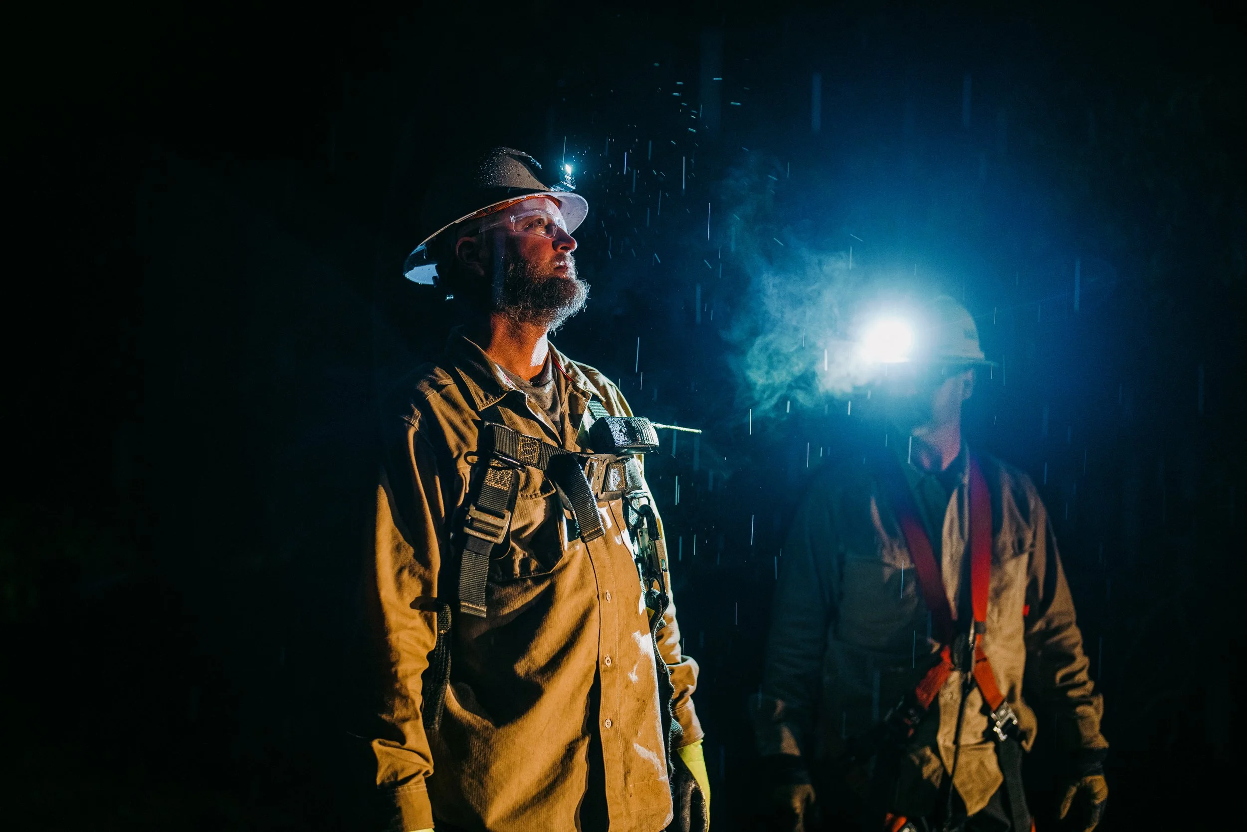 Two construction workers wearing hard hats and safety gear working at night in the rain.