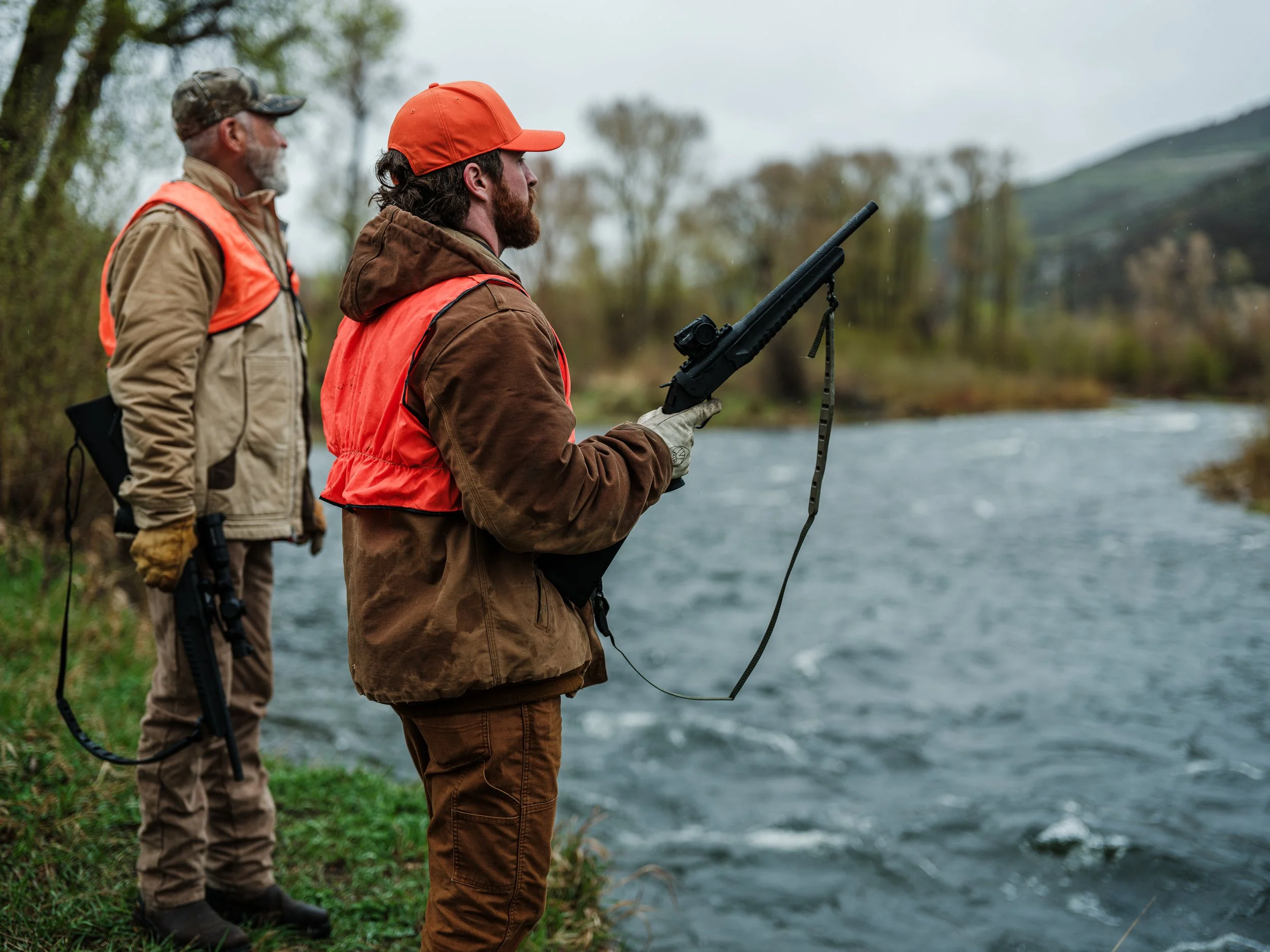 Two hunters in orange vests holding rifles by a river in a wooded area.