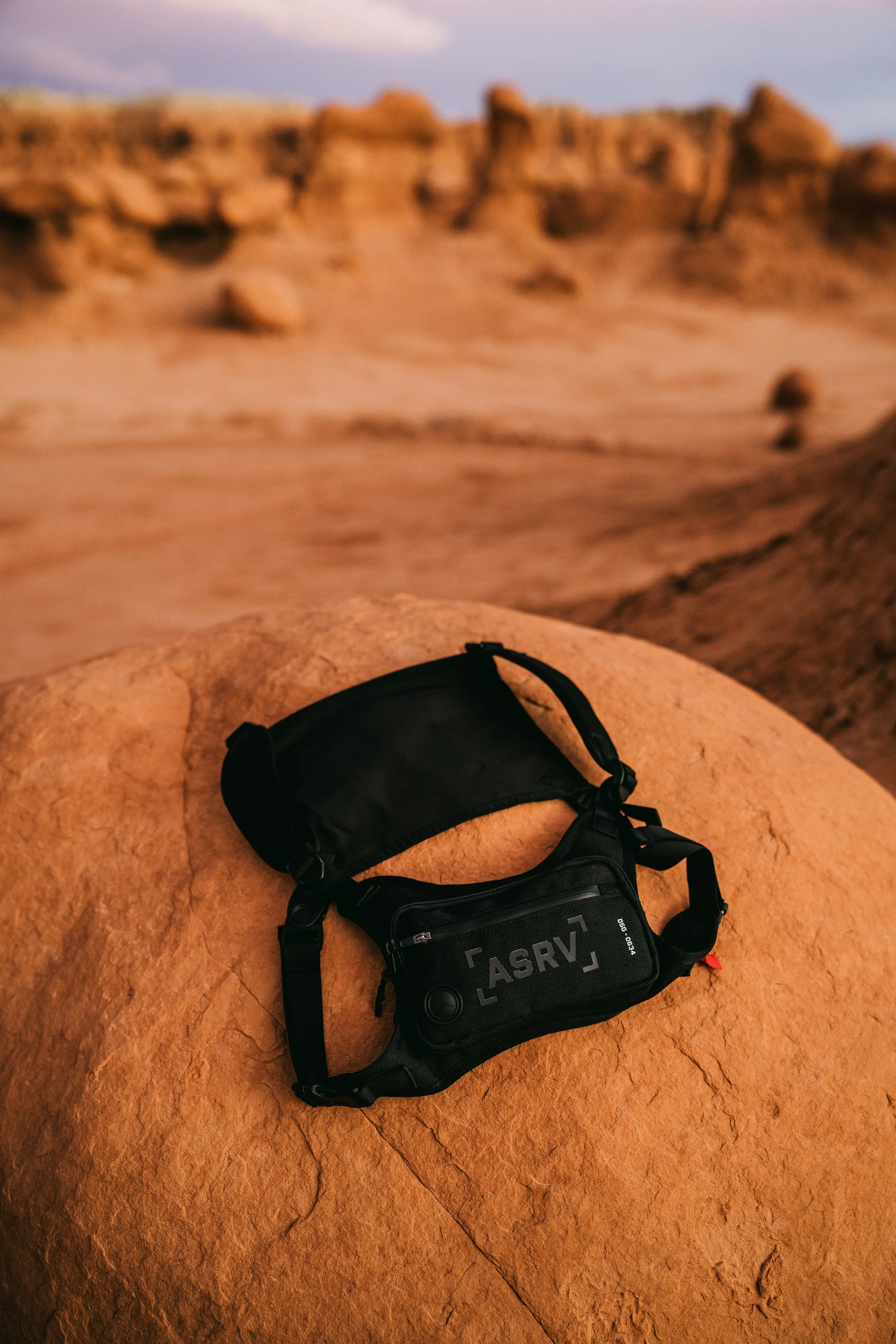 A black tactical chest bag on a rock in a desert landscape during sunset.