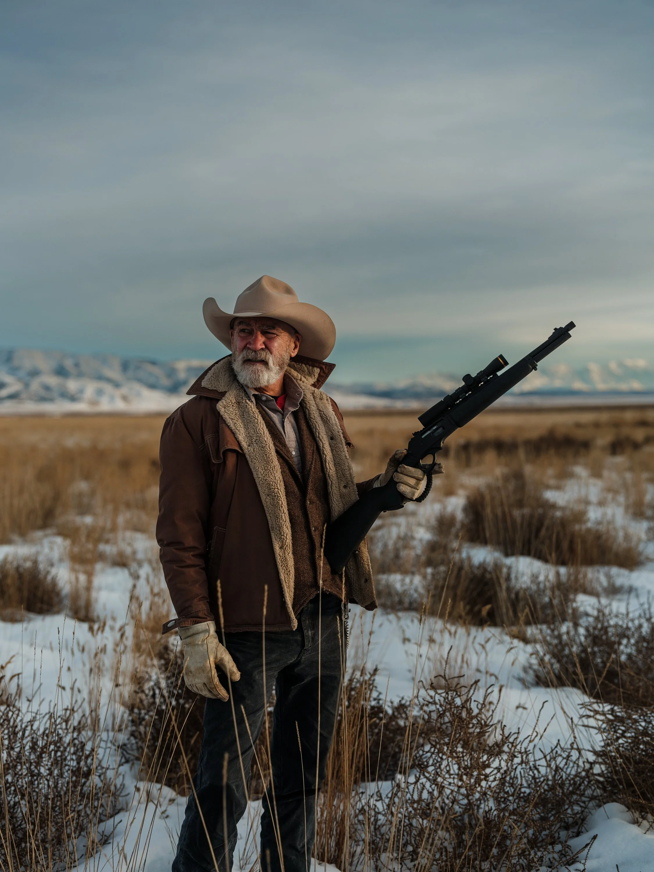 Older man in a cowboy hat and winter jacket holding a rifle, standing in a snowy field with mountains in the background.