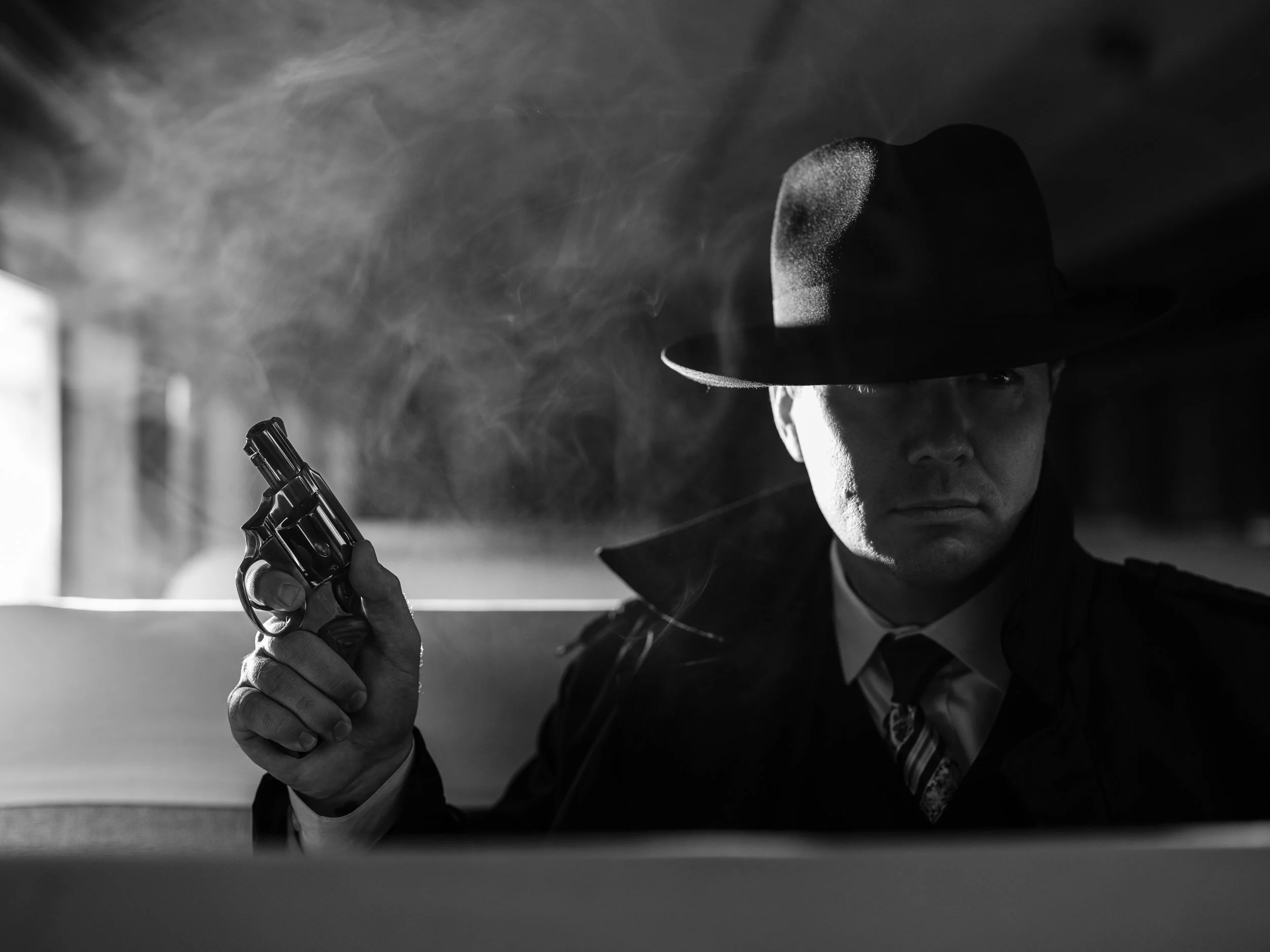 Black and white image of a man in a fedora and trench coat holding a revolver, with smoke around him, in a noir-style setting.