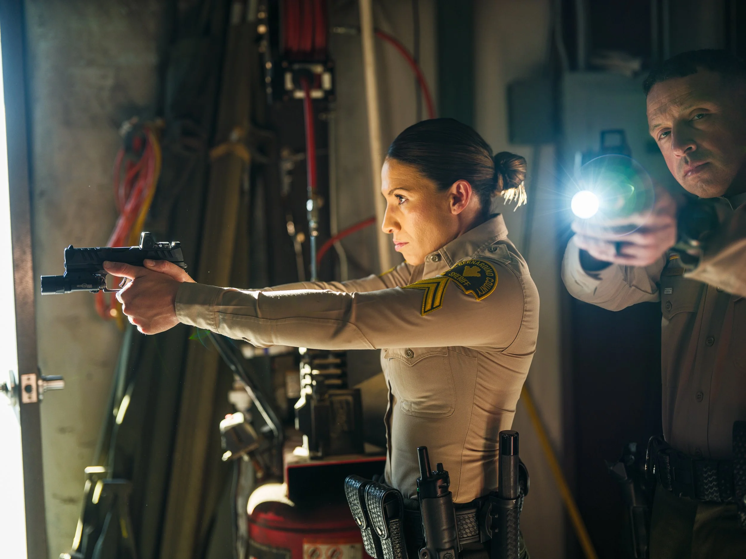 Two uniformed sheriffs, one aiming a gun with a mounted flashlight, the other pointing a flashlight, standing in a dimly lit room.