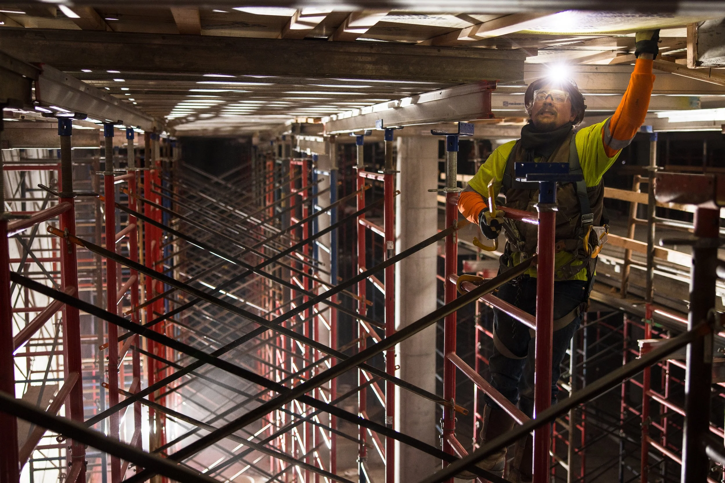 Construction worker on scaffolding with a headlamp in an industrial setting, surrounded by metal beams and wooden planks.