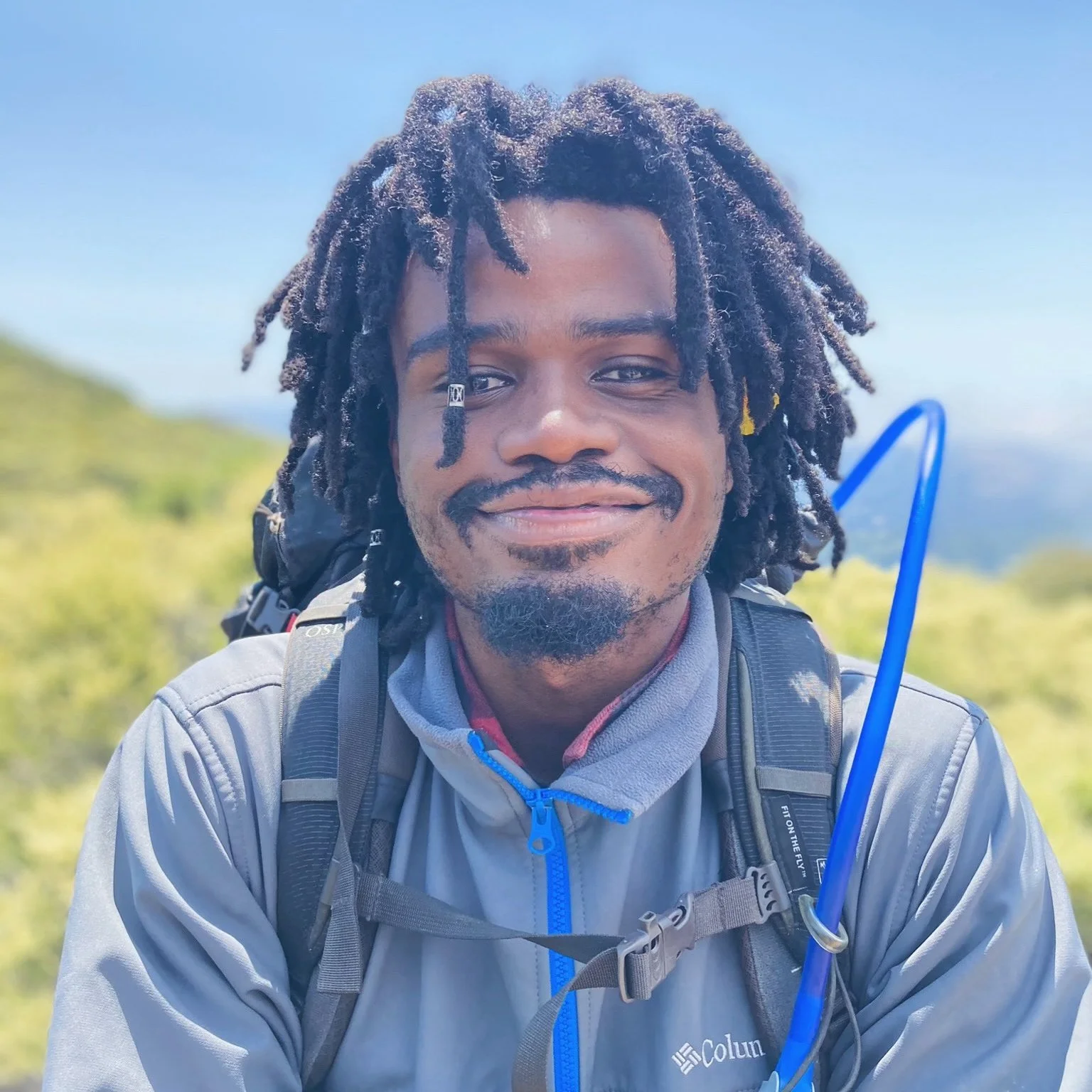 A man with dreadlocks and facial hair, wearing a black shirt and a red backpack, looking surprised or excited outdoors on a sunny day.