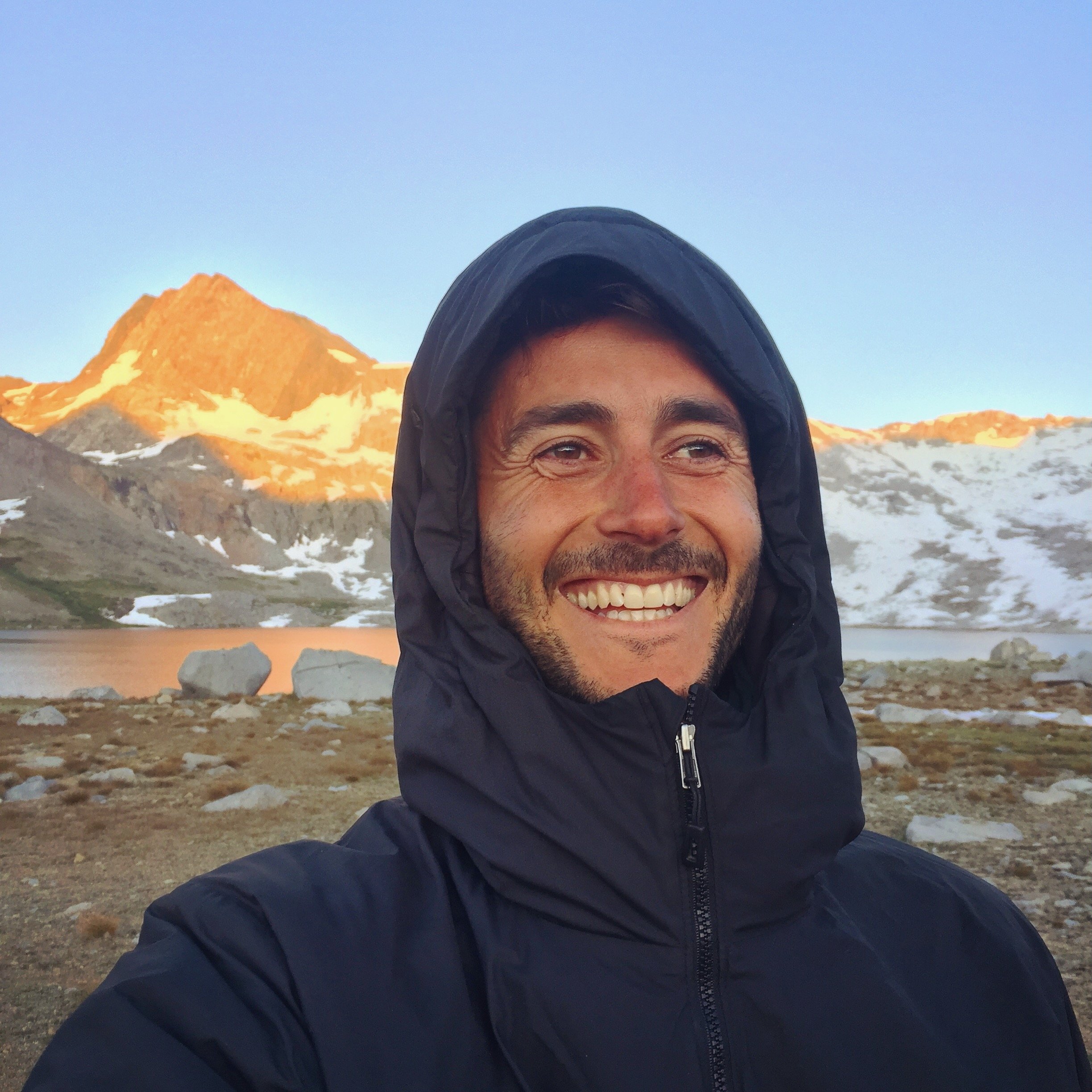 A man outdoors in front of a mountain, smiling, with rocky background.