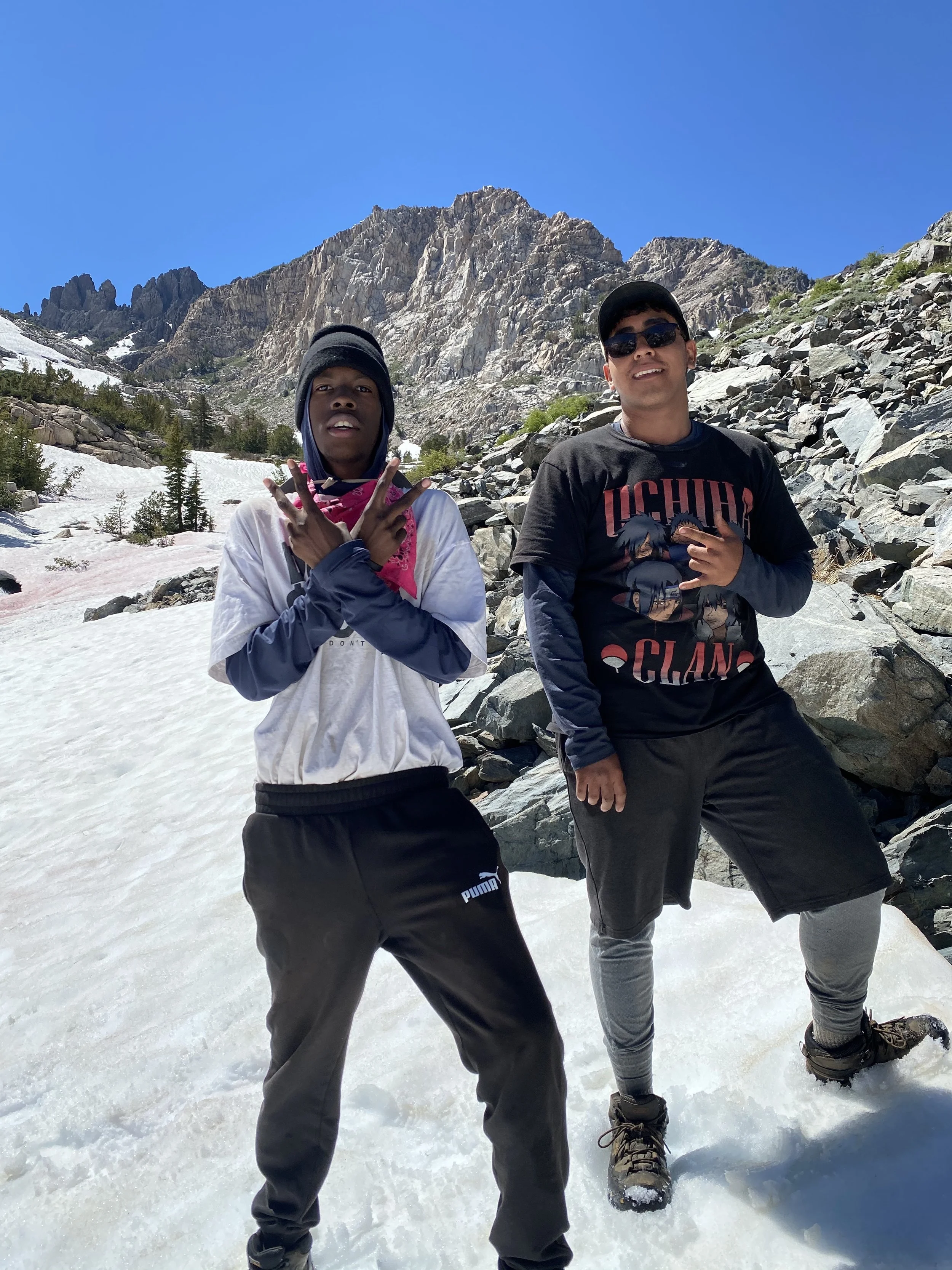 Two young men posing on snow-covered ground in a mountainous landscape under a clear blue sky. One is making a peace sign with both hands, and the other is holding his hand over his chest. They are dressed in casual outdoor clothing, including hoodies, sweatpants, and hiking boots.