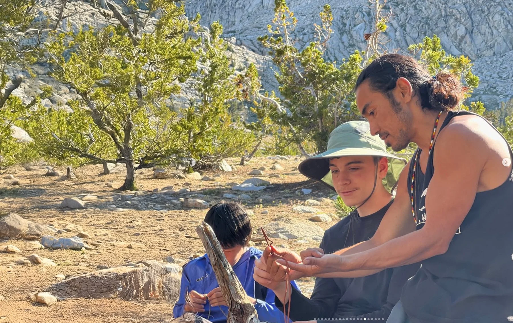 Three people sitting outdoors in a rocky and grassy area with green trees and a rugged mountain behind them. One man with long hair and a sleeveless shirt shows something to a young boy wearing a hat, while another young girl with dark hair looks on.