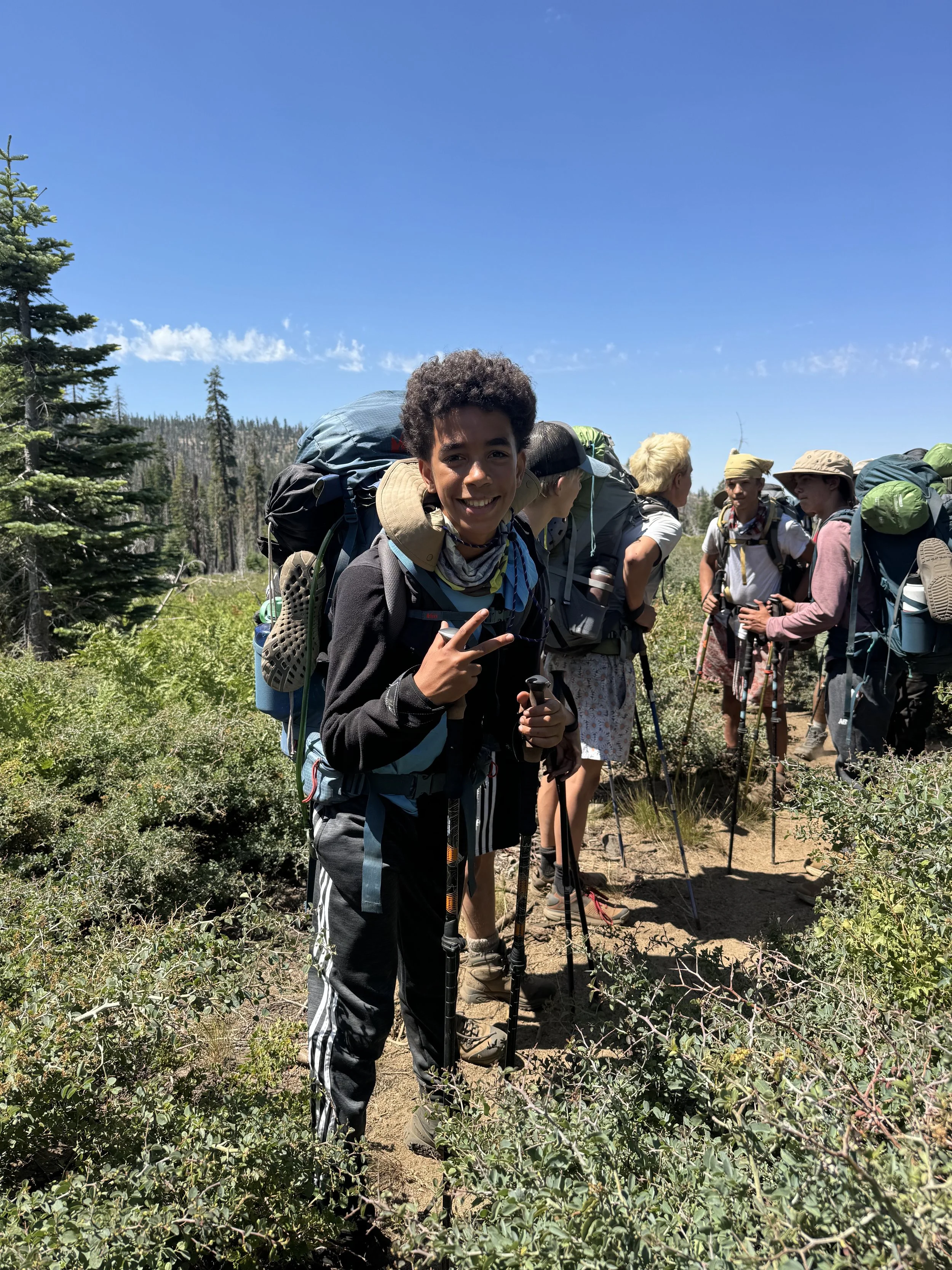 Group of people hiking on a trail in a forested area under a clear blue sky, with one person smiling and making a peace sign in the foreground.