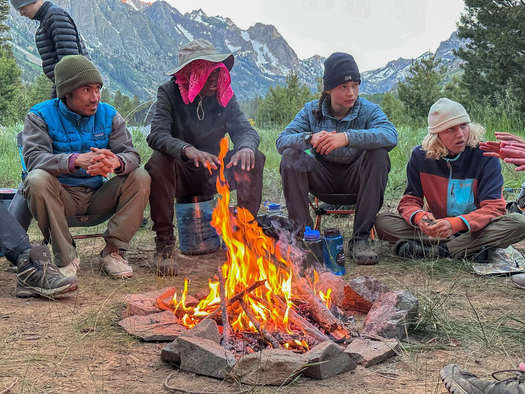 A group of people camping outdoors around a campfire with mountains and trees in the background. They are sitting on the ground or small chairs, wearing outdoor clothing and hats.