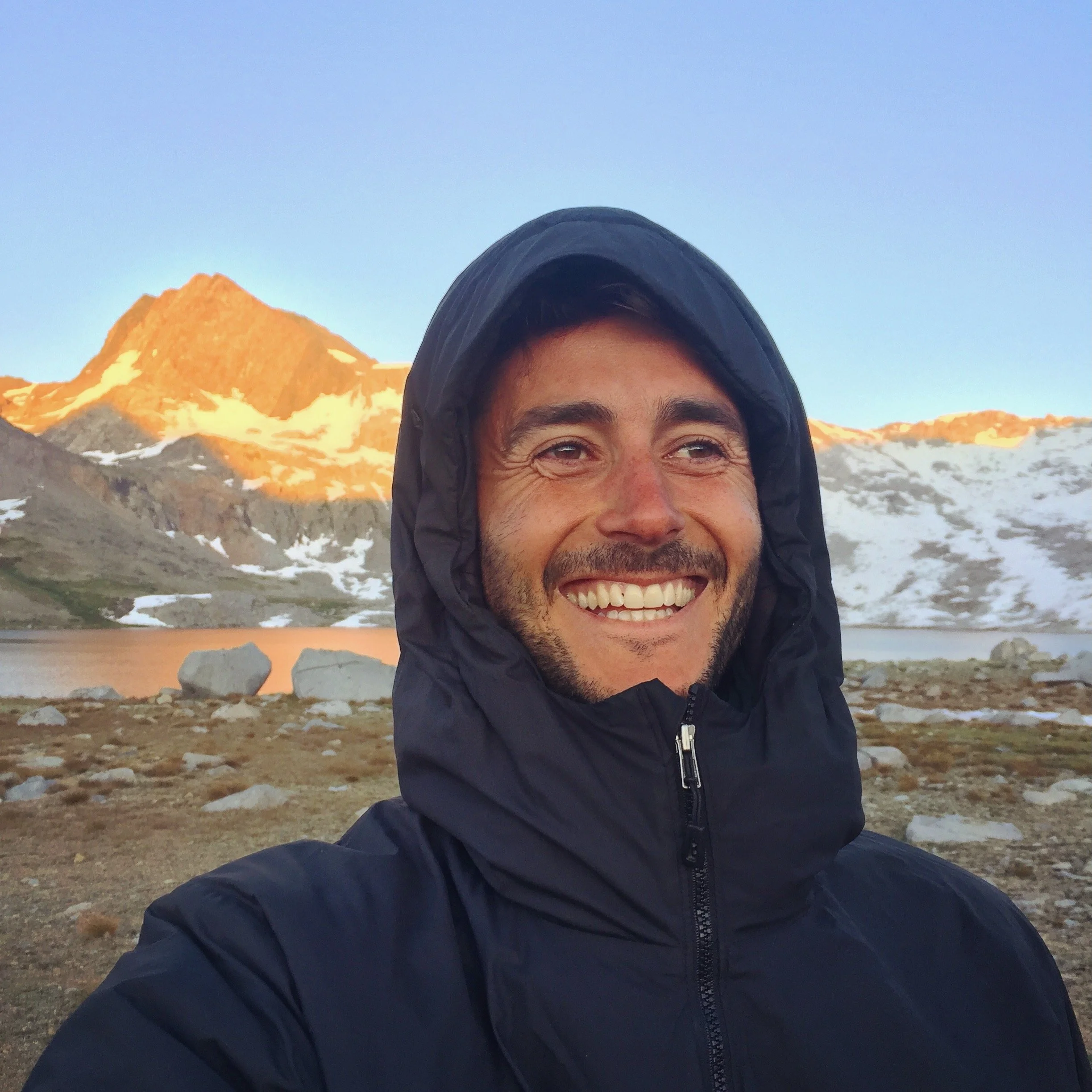 A man smiling and sitting outdoors while holding a smartphone, with rocks in the background.
