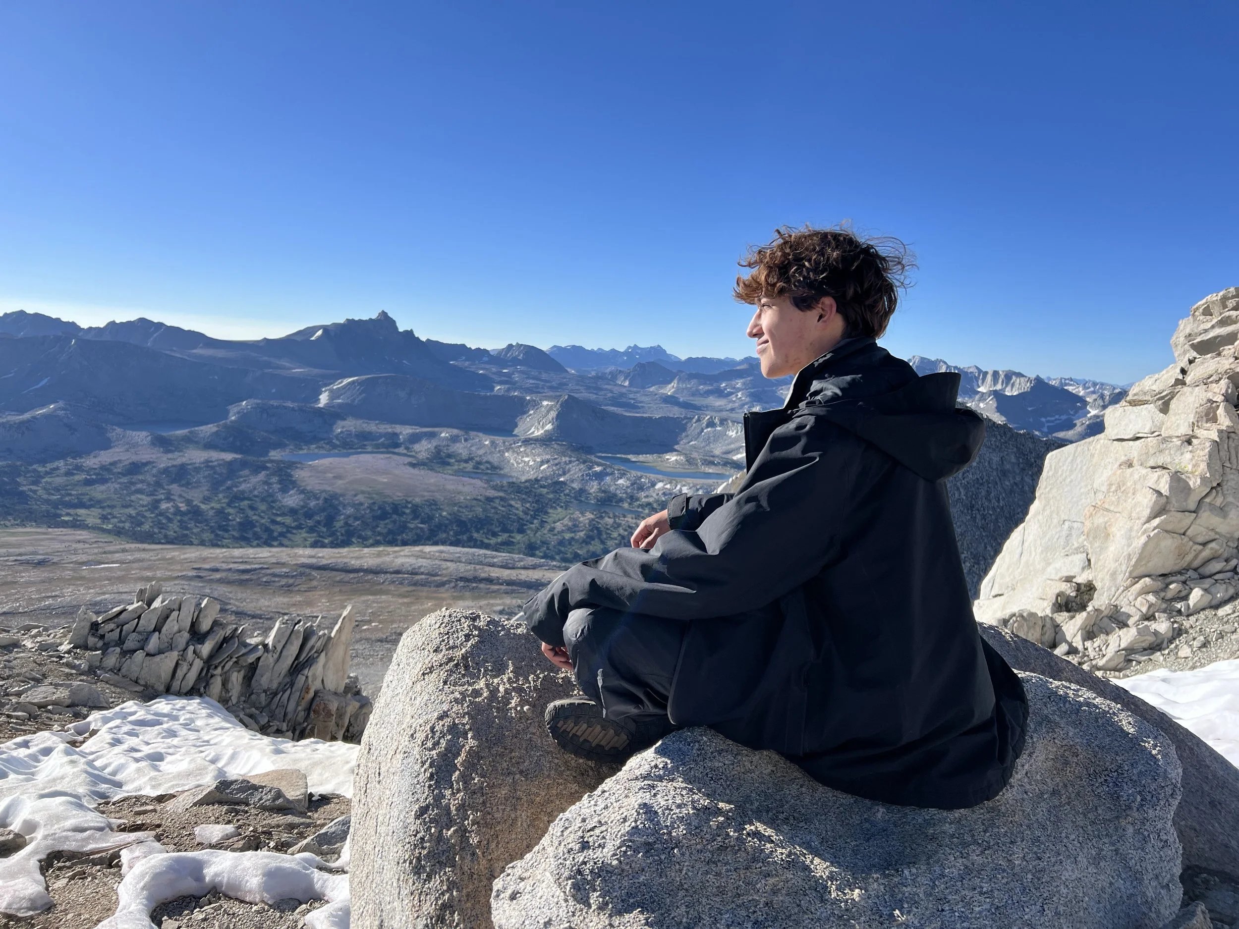 A person sitting on a large rock on a mountain viewpoint, looking into the distance at snowy mountain peaks and valleys under a clear blue sky.