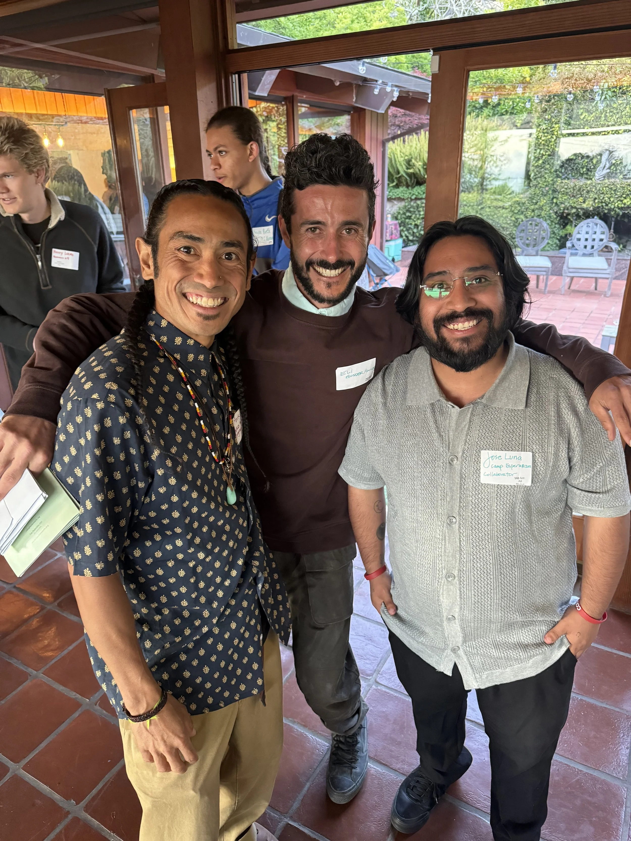 Three men standing together indoors with open doors and windows behind them, smiling for a group photo at a social gathering.