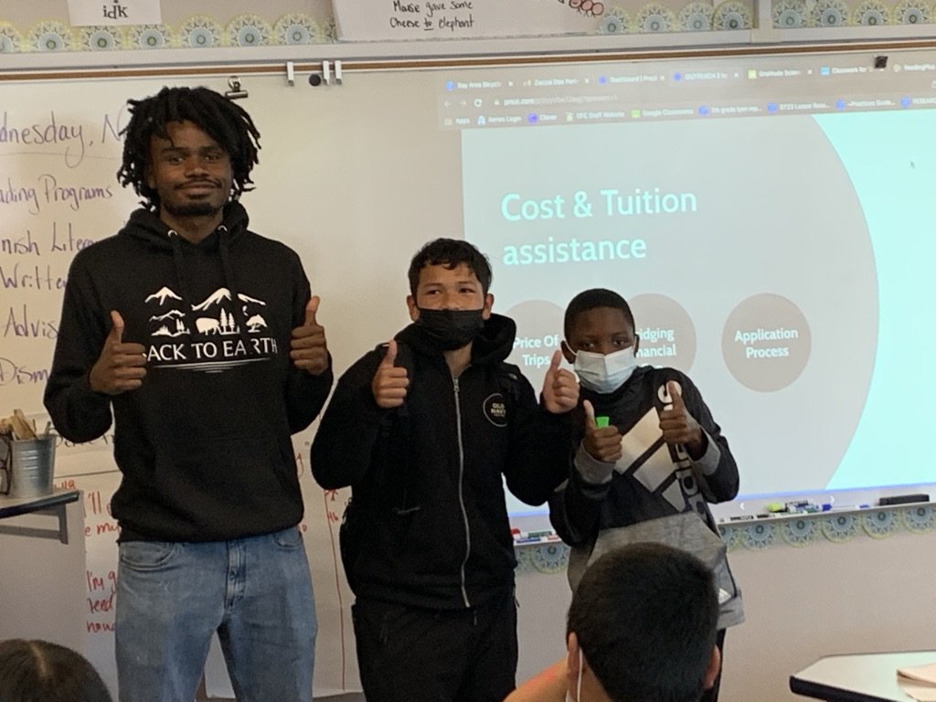 A male teacher and two boys wearing face masks, standing in front of a classroom whiteboard and screen showing a PowerPoint slide discussing cost and tuition assistance, all giving thumbs-up.