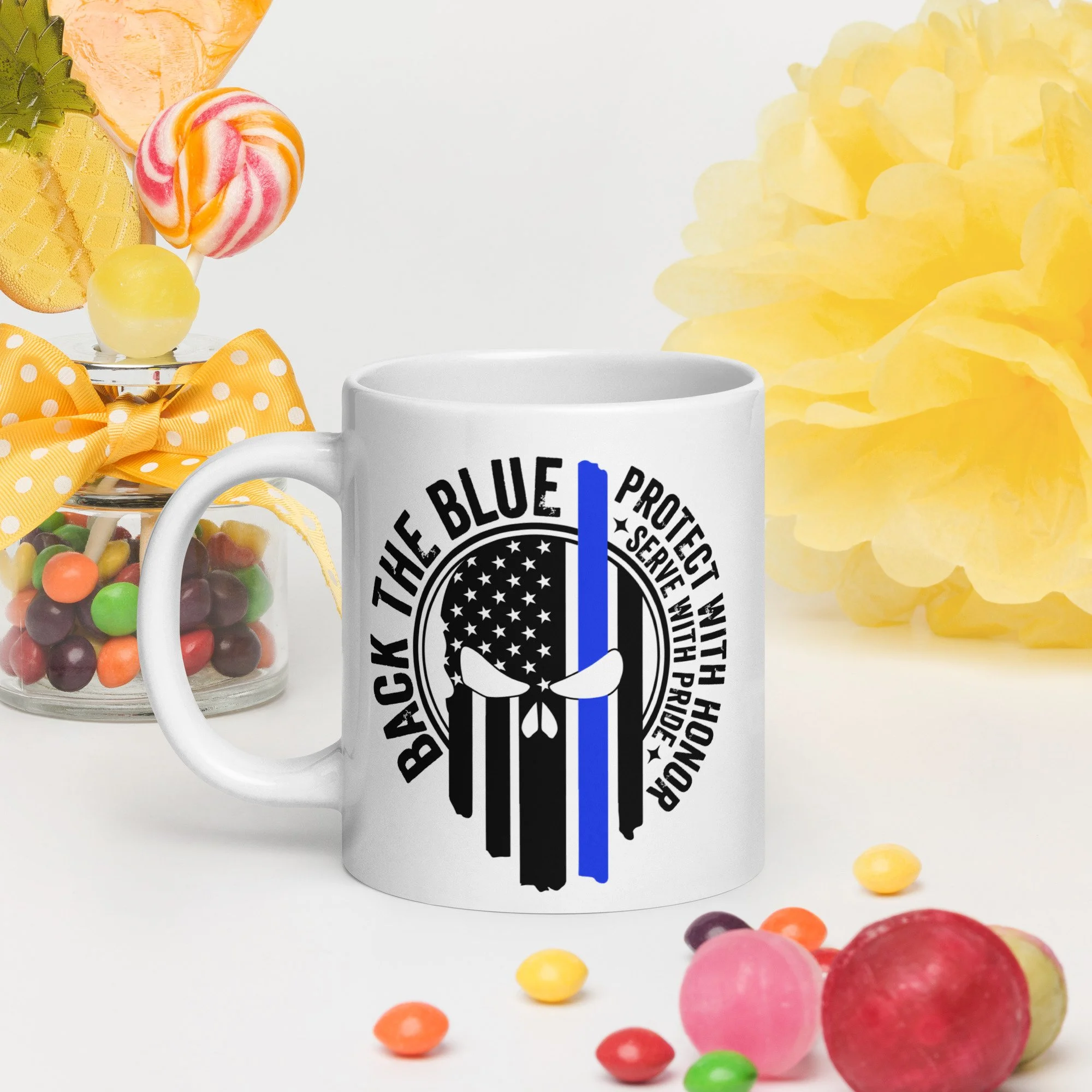 A white mug with a logo and Blue Line "Back the Blue", surrounded by colorful candies and yellow paper flowers.