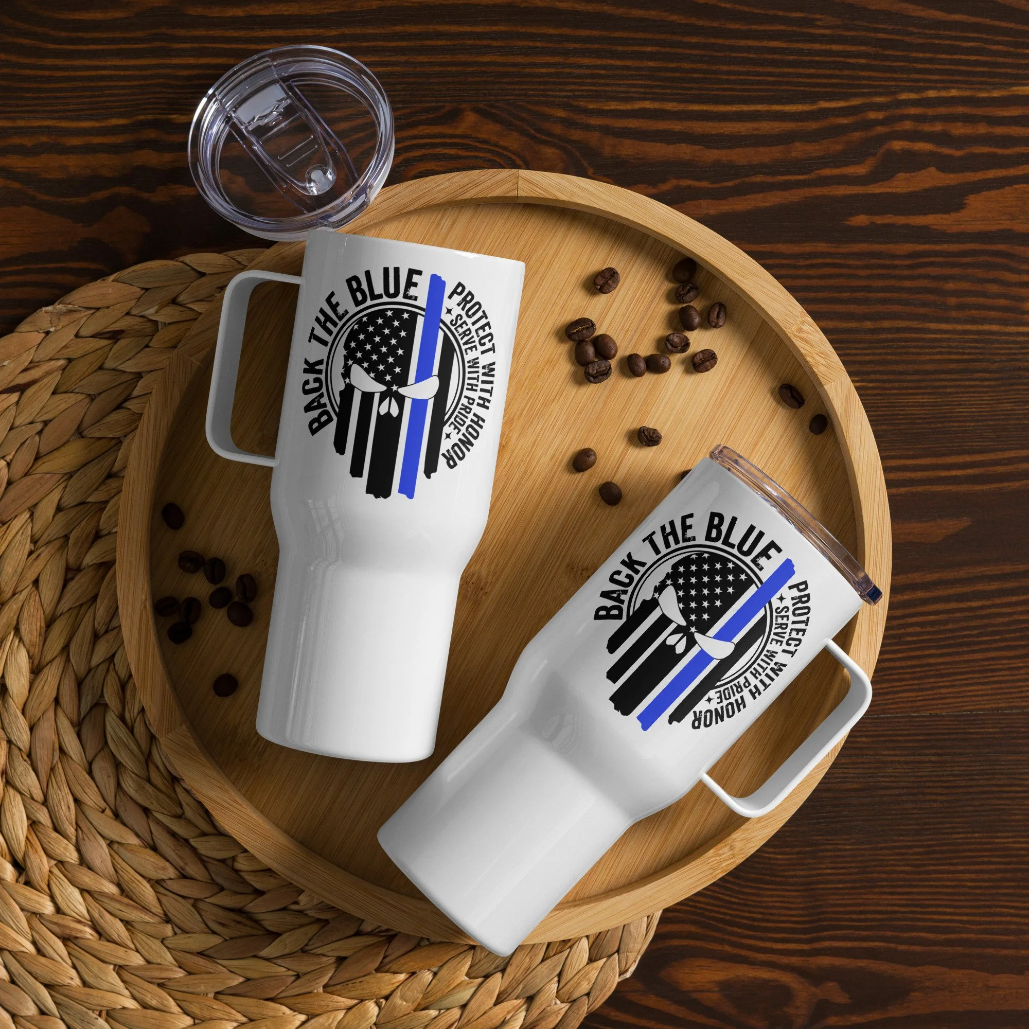 A white mug with black text that reads "Back the Blue" and a blue line, resting on a round wooden tray with scattered coffee beans. Next to the mug is a clear plastic cup and a white rectangular object on a woven placemat on a wooden table.