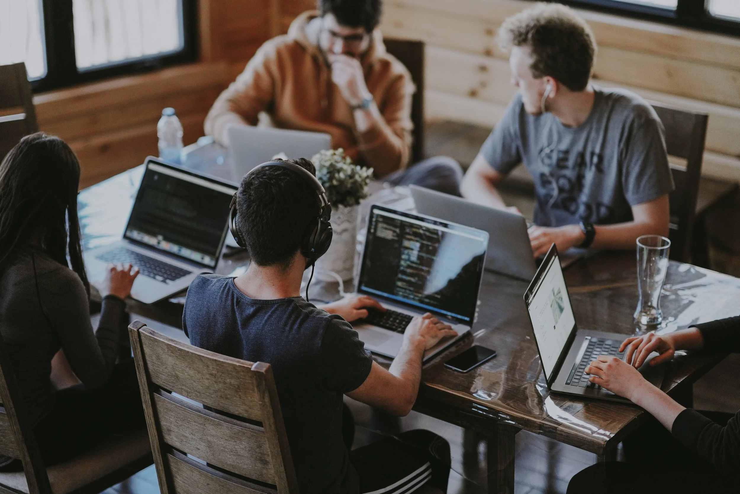 A group of five young adults working together around a wooden table with laptops, some wearing headphones, in a cozy room with wooden walls and large windows.