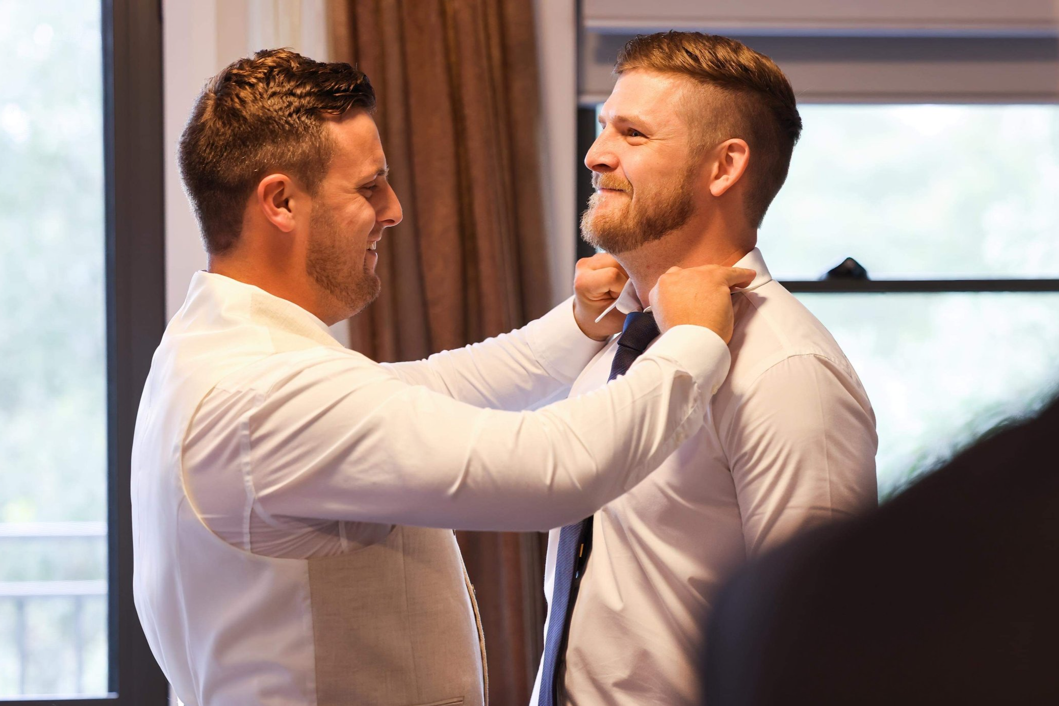 Two men, dressed in white shirts, smiling and helping each other adjust ties, indoors with windows in the background.